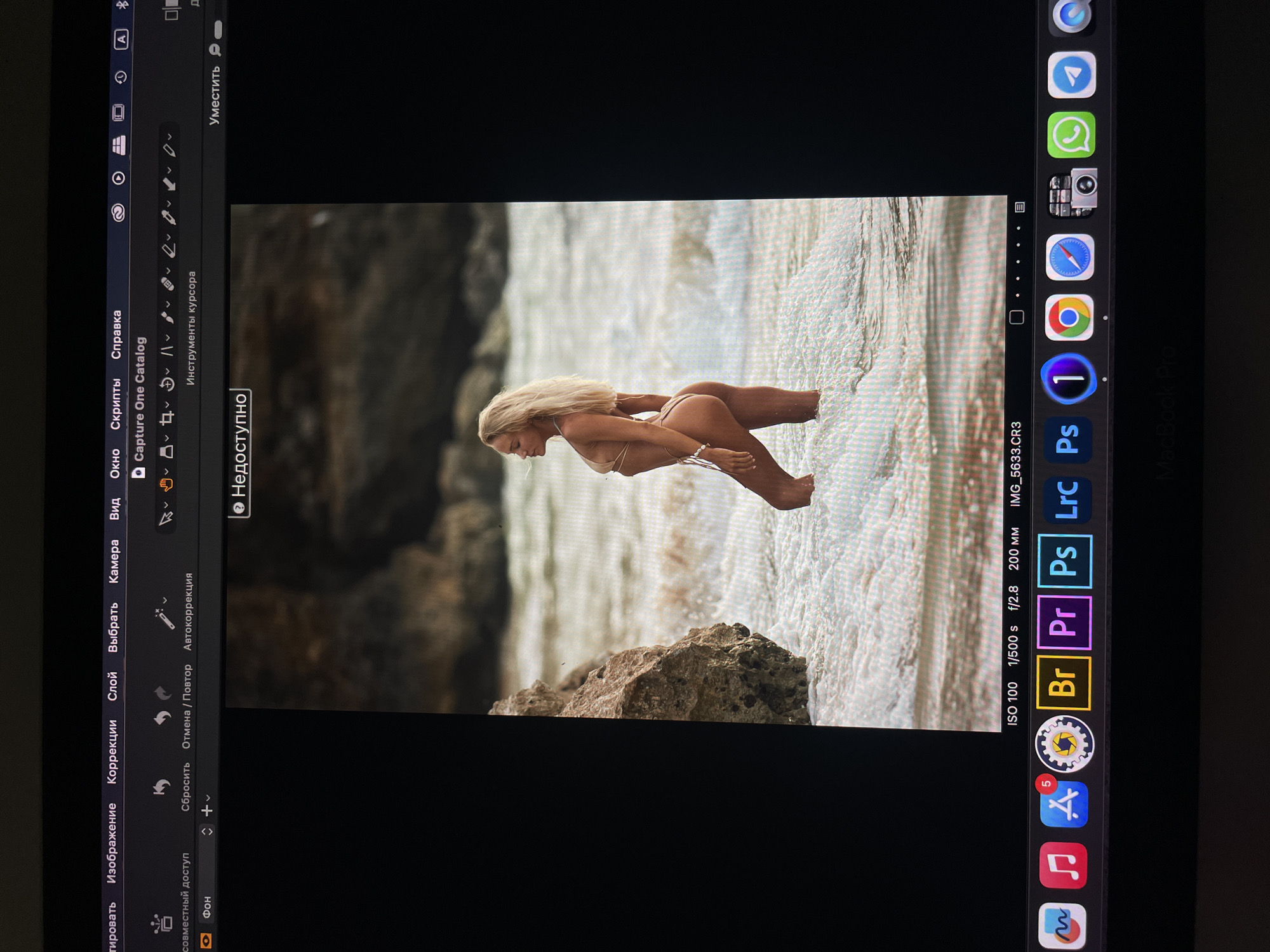 A photograph of a computer screen depicts a woman in a bikini standing in water near a rocky outcrop. The computer interface suggests photo editing software is open. The taskbar shows various applications including Adobe Photoshop, Premiere Pro, and Bridge. The camera settings are displayed as ISO 100, 1/500 s, f/2.8, 200 mm. The location, Kazan, Russia, is not evident in the image.
