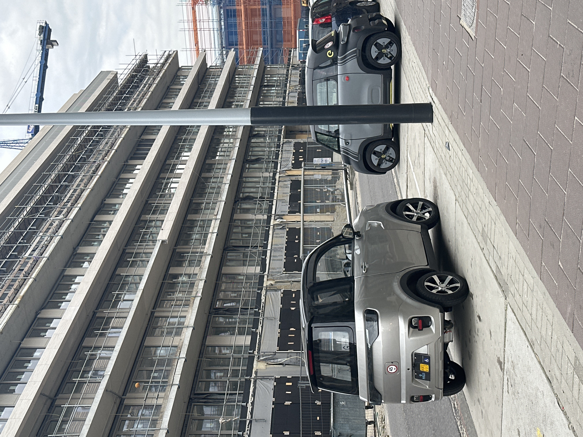 The image captures a street scene in Amsterdam, Netherlands. The view is dominated by a tall, aging office building undergoing extensive renovations, with scaffolding covering much of its facade. Parked along the curb are several compact, electric vehicles, possibly Citroën Ami models, including a silver one and a grey one. The pavement is a mix of grey stone and red brick. A light pole extends from the bottom left corner diagonally across the frame. A crane is visible in the background. The sky is overcast. There are no people visible in the immediate foreground. The overall impression is one of urban renewal alongside contemporary, eco-conscious transport solutions.
