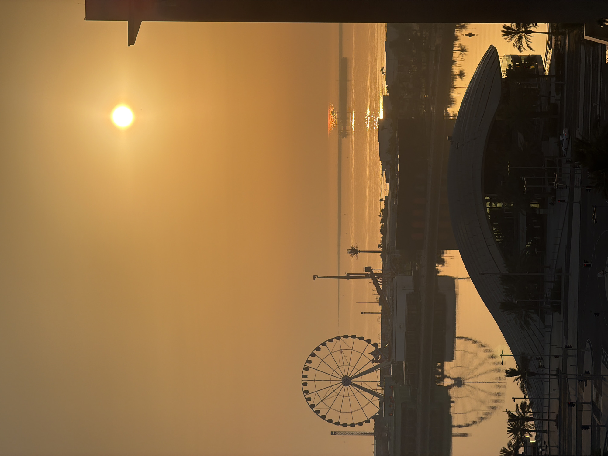 A Ferris wheel and other amusement park rides are silhouetted against a hazy orange sky at sunrise. The sun, a bright orb, is positioned in the upper right quadrant of the image, casting a warm glow over the scene. The Ferris wheel, on the left, is a prominent feature, its spokes clearly defined. In the foreground, a modern building with a curved roof and palm trees are visible. The water in the middle ground reflects the sky and the Ferris wheel. A ship is visible in the distance, its reflection shimmering on the water. The overall scene suggests a coastal city with an amusement park.