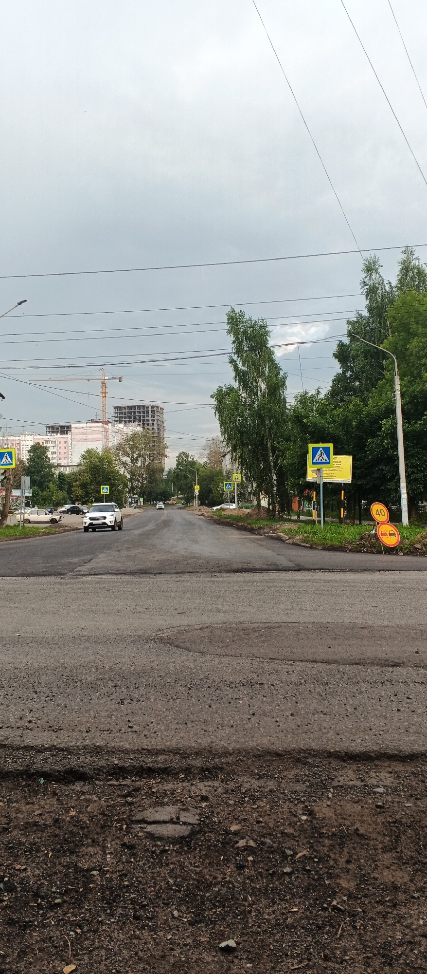 The image shows a street scene in Izhevsk, Russia, on a cloudy day. The foreground is dominated by a rough asphalt road with visible patches and unevenness, indicating recent or ongoing road work. In the middle ground, the road curves slightly and continues towards a residential area. On the left side of the road, a white car is driving, and further down, several other cars are parked or moving. There are tall trees lining both sides of the street. Several yellow pedestrian crossing signs with a black pedestrian symbol are visible along the road. In the background, a large, multi-story building under construction with a yellow construction crane is prominent. Overhead, power lines crisscross the sky. On the right side of the road, there are two circular traffic signs, one indicating a speed limit of 40 and another with an image of cars, suggesting a road hazard or lane closure. The overall impression is of an urban environment undergoing development or repair, with a slightly overcast sky contributing to a subdued atmosphere.
