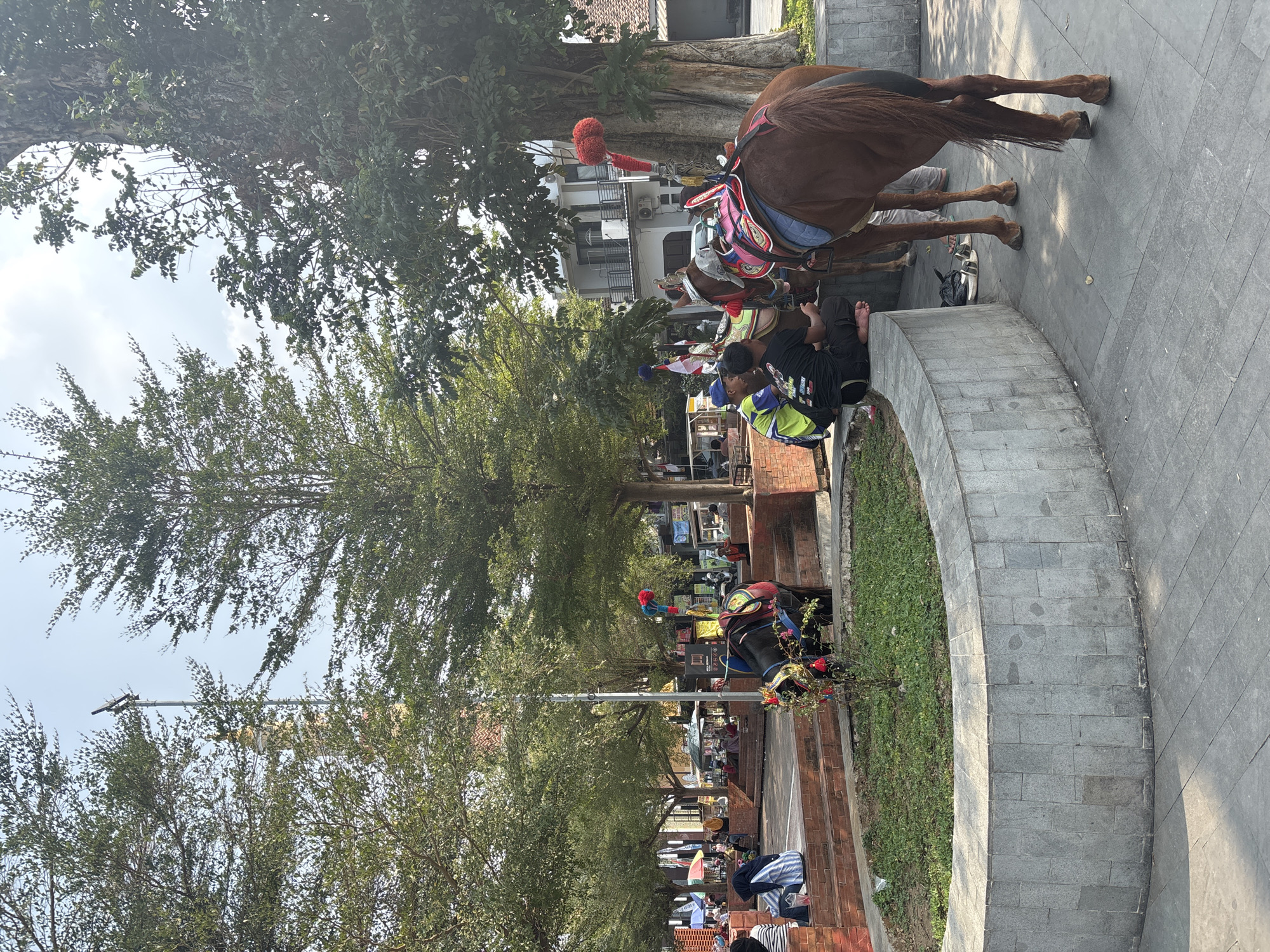 A brown horse, adorned with colorful saddle blankets and decorative tassels, stands on a paved area. Two men, one wearing a blue cap and a yellow and green shirt, and the other in a black t-shirt, sit on a stone wall nearby, facing away from the horse. In the background, several other horses are visible, some with riders. There are also various market stalls and awnings, with people browsing. Trees with lush green foliage dot the scene, and a tall, slender tower, possibly a minaret, can be seen rising above the trees. The area appears to be a public square or market, likely in Majalengka, Indonesia, given the traditional horse-drawn carriage setup, known as 'andong' or 'delman', commonly found in tourist areas of Indonesia. The lighting suggests it is daytime, with sunlight casting shadows on the ground.