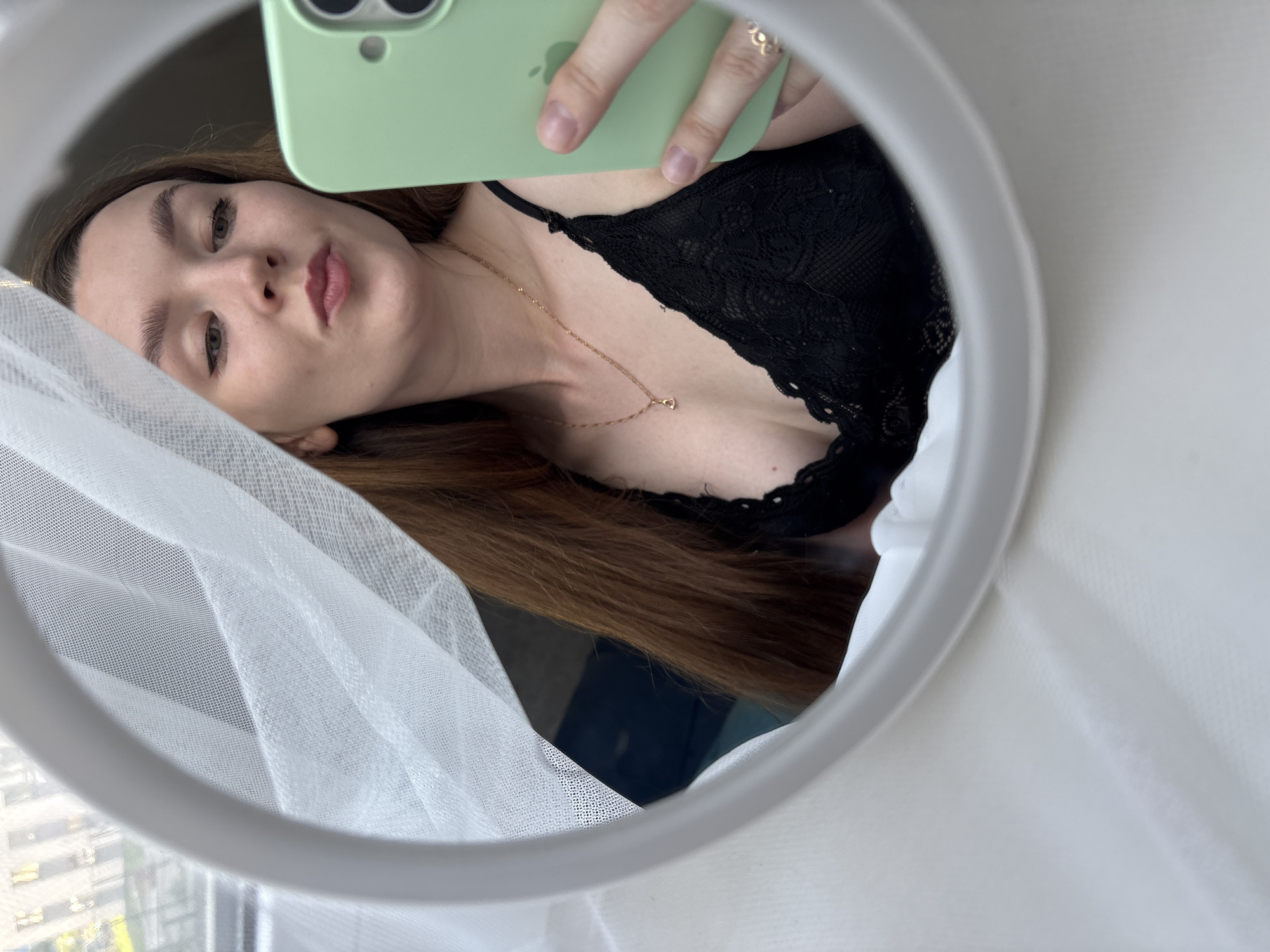 A young woman with long, auburn hair is reflected in a mirror, taking a selfie. She is wearing a black lace bralette and a delicate gold necklace with a heart pendant. Her left hand holds a light green smartphone, and a gold ring with a heart detail is visible on her right hand. A sheer white curtain partially obscures her hair and the background, which appears to be a window overlooking buildings. The setting suggests an indoor environment, likely a bedroom or living area.