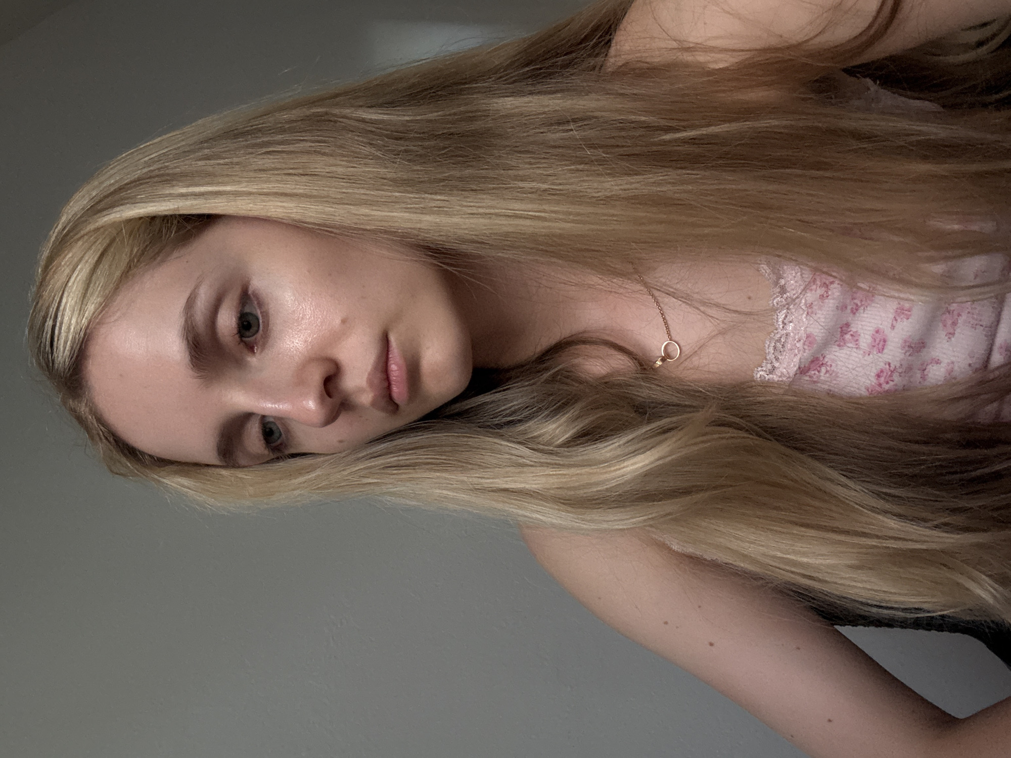 This is a portrait photograph of a young woman with long, blonde hair. She is lying down, with her head resting on her arm. Her eyes are open and she is looking slightly away from the camera. She is wearing a pink tank top with a floral pattern and a lace trim. A delicate gold necklace with a circular pendant is visible around her neck. The background is a plain, light gray wall. The lighting suggests an indoor setting, possibly in the afternoon. The overall mood is relaxed and introspective. The photograph appears to be taken in Miami, United States of America. There is no visible text in the image.