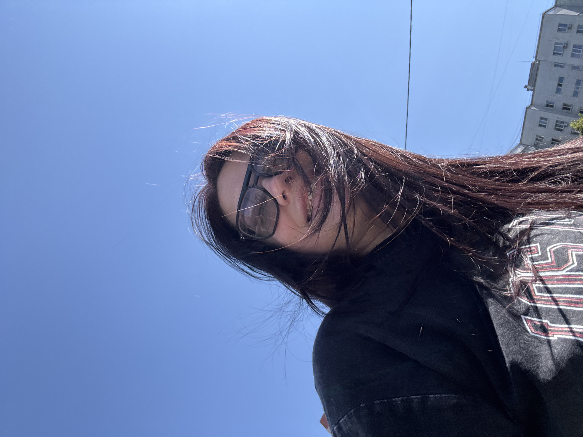 A low-angle selfie shows a woman with long, dark hair blowing in the wind against a bright blue sky. She is wearing large, dark-rimmed sunglasses and a black sweatshirt with lettering. Her mouth is slightly open, revealing teeth. In the background, a light-colored multi-story building with visible windows and a power line are present. The scene suggests an outdoor urban environment. The location is Tolyatti, Russia.