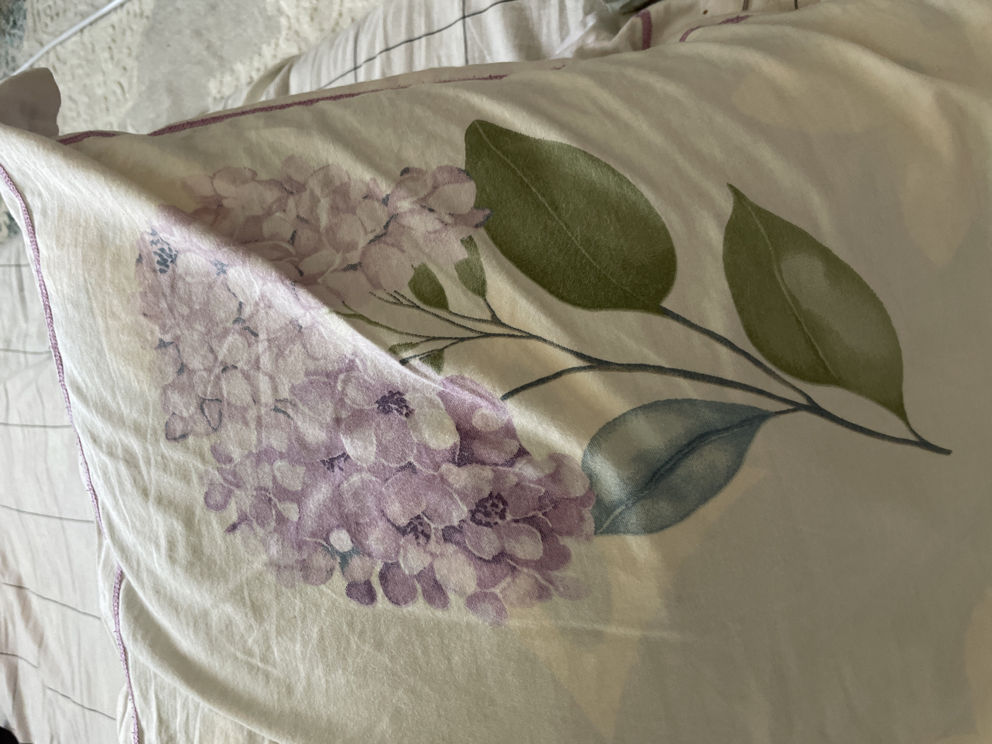 The image shows a close-up of a pillow with a floral pattern, likely in a bedroom. The pillow is light-colored with a print of purple hydrangeas and green leaves. The fabric is slightly creased, suggesting it's in use. The background shows bedding with thin lines, possibly a bedsheet or duvet cover. The lighting appears to be natural, coming from an unseen source, casting subtle shadows on the pillow. There are no people or animals visible in the image. There is no discernible context to place this image in a specific city or country, including Чат-Көл, Kyrgyzstan.