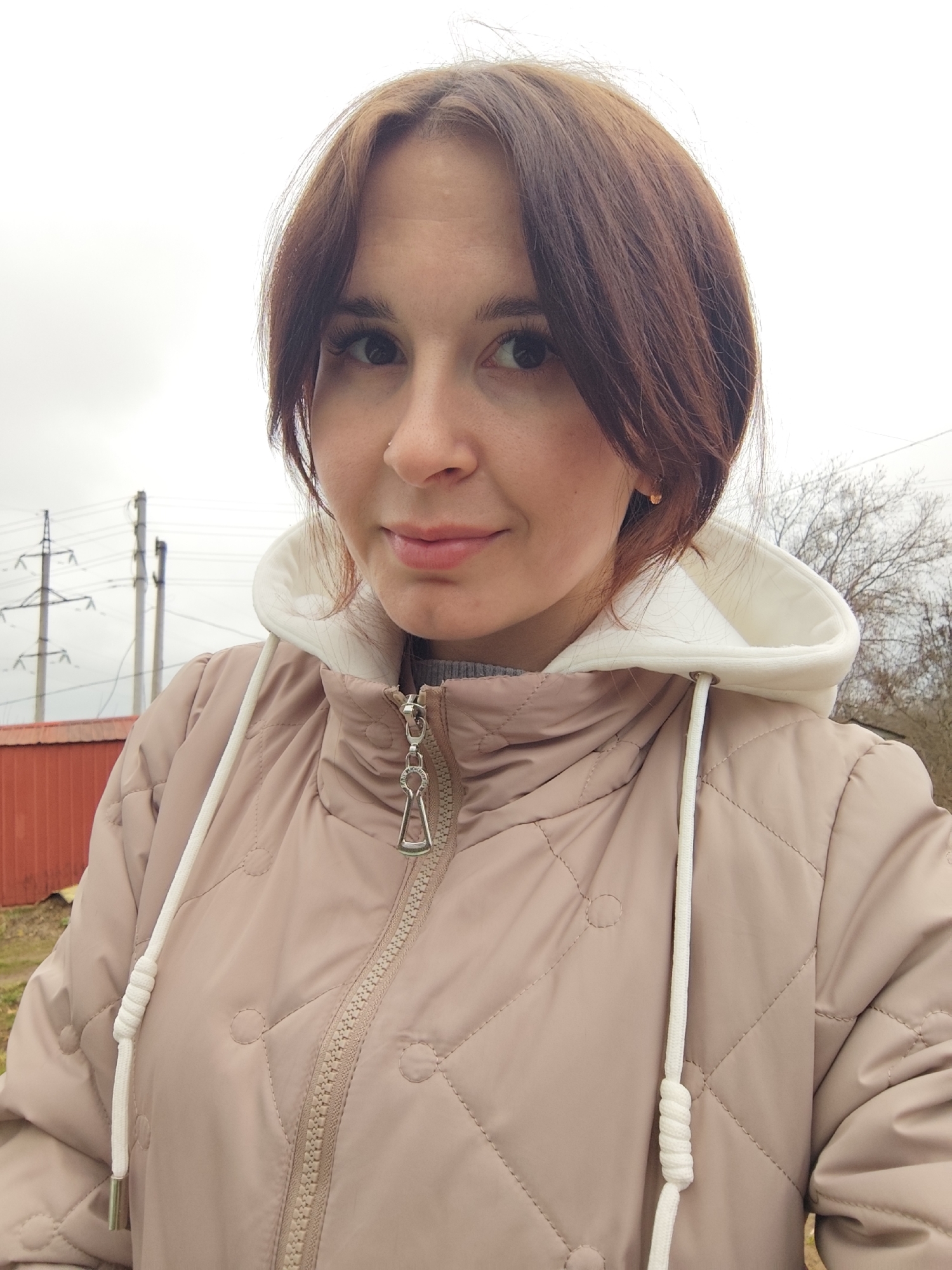 This is an outdoor close-up portrait of a woman, likely a selfie, taken in Фоки, Russia. The woman has brown hair, dark eyes, and a small nose piercing on her right nostril. She is looking directly at the camera with a neutral to slightly pleasant expression. She is wearing a light beige or pale brown quilted jacket with a white inner hooded layer. The jacket features a silver zipper with a unique pull designed like a stylized keyhole or figure-eight, and white drawstrings for the hood.
The background is overcast, indicating a dull or cloudy day. On the left side of the frame, several power lines and utility poles are visible, along with a partial view of a red building or structure. On the right side, bare tree branches can be seen against the sky, suggesting a cold season such as late autumn, winter, or early spring. No discernible text is visible in the image.