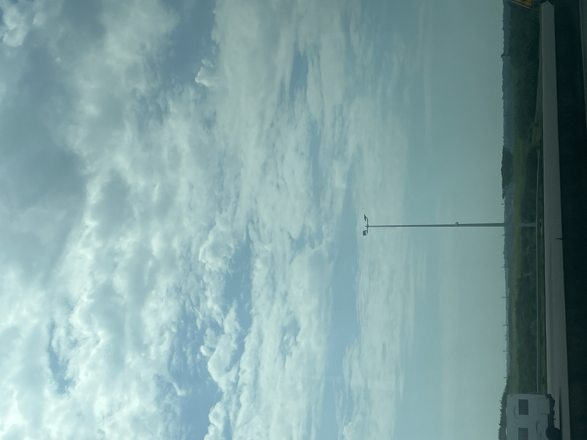 The image depicts a landscape view with a wide expanse of sky filled with clouds. In the foreground, a dark road or highway runs horizontally across the bottom of the frame. To the left, a white caravan with visible windows is parked on the side of the road. Further in the distance, a tall lamppost with a double light fixture stands prominently in the middle ground. Beyond the lamppost, a green, treed landscape stretches to the horizon under the cloudy sky. The overall scene suggests an outdoor, possibly rural or semi-rural, environment. There is no indication of the city of Thun-l'Évêque, France within this image.