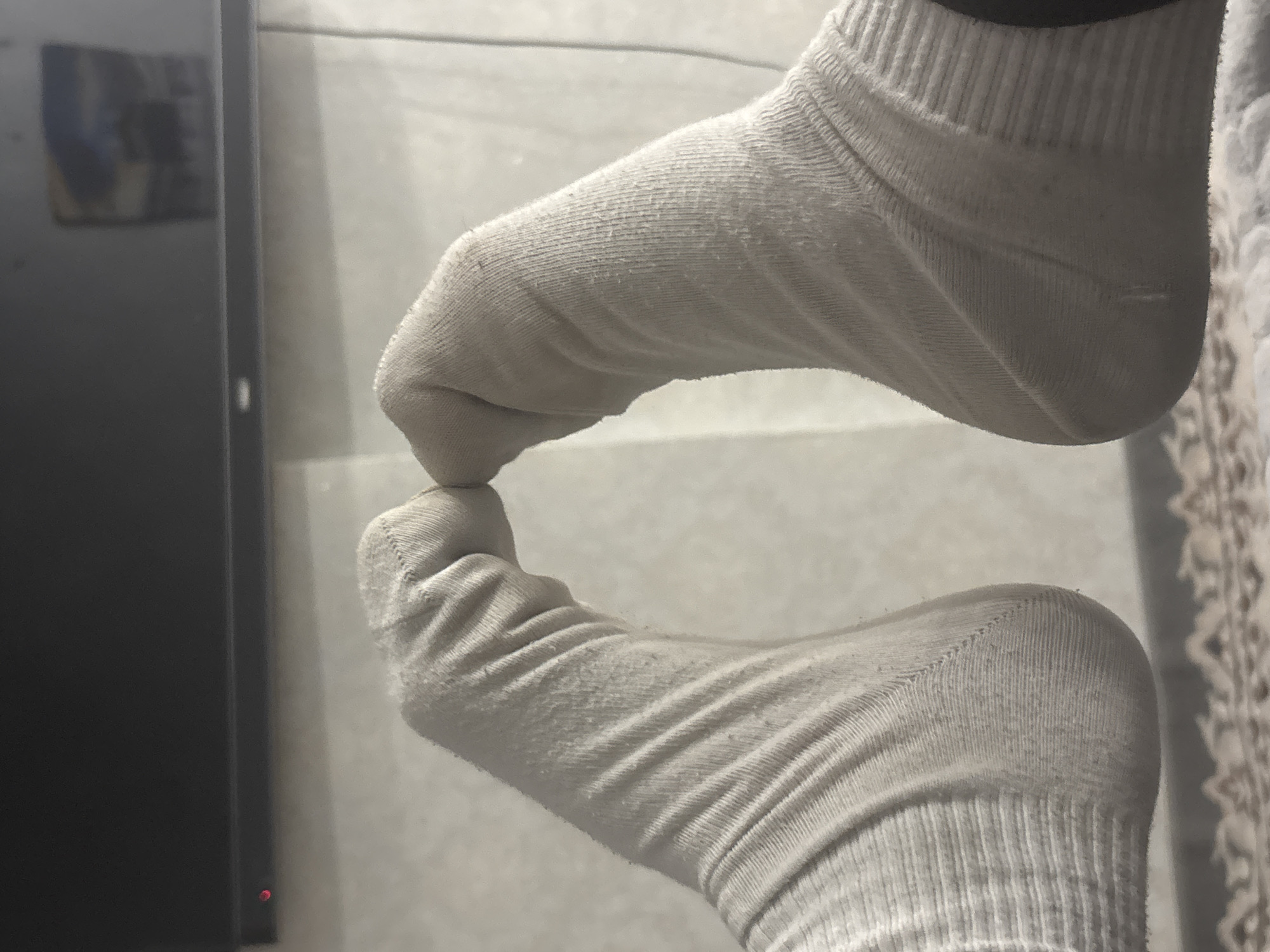 The image shows two feet wearing white socks, placed heel-to-heel, with the toes touching. The socks are ribbed and appear to be slightly worn. The background is out of focus, but appears to be an indoor setting with a dark object, possibly a television, in the upper portion of the image and a tiled wall behind the feet. There is no information in the image that can place this scene in Vaganovo, Russia.