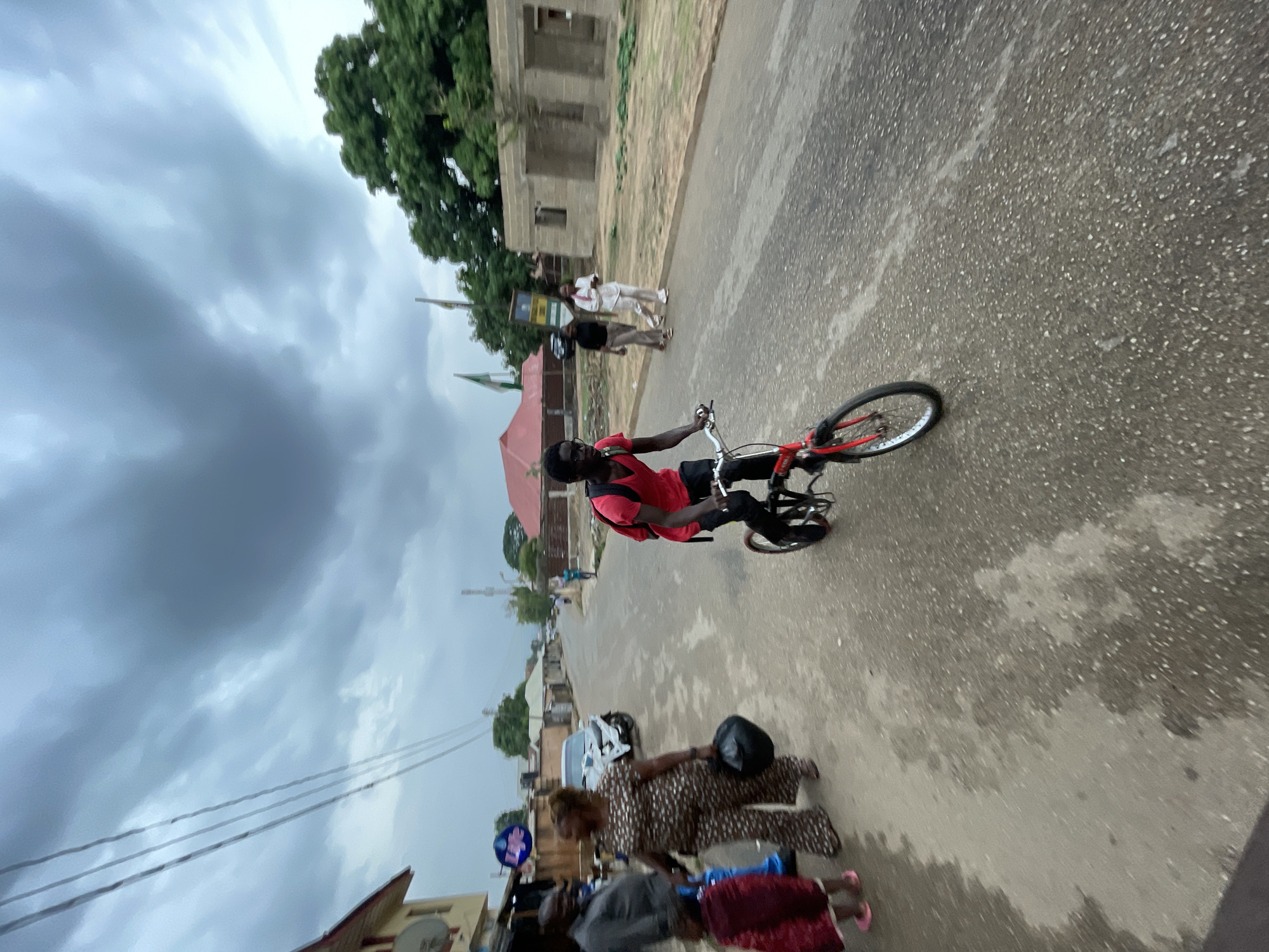 The image captures a street scene in Jos, Nigeria. A young man wearing a red shirt and black pants is riding a red bicycle on a paved road. There are people walking on the sidewalk and along the street. In the background are buildings, trees, and a cloudy sky.