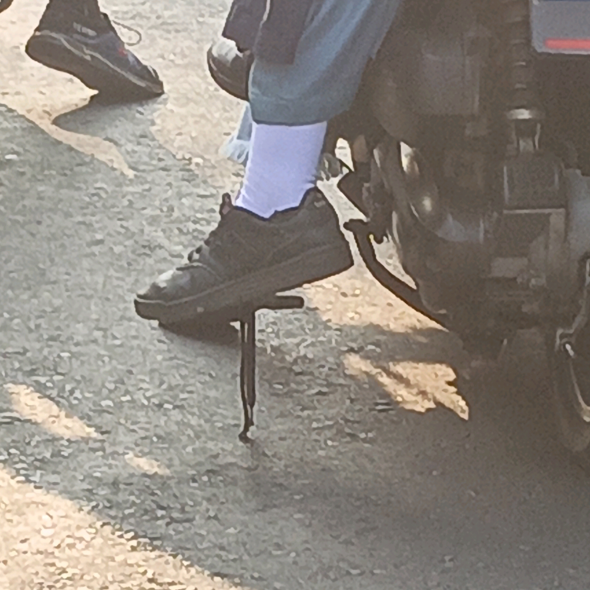 The image depicts a low-angle, close-up shot of a person's feet and lower legs. One foot is clad in a dark-colored sneaker with white socks visible above it. This foot rests on a motorcycle kickstand, which is extended and in contact with the ground. The ground appears to be paved with asphalt or gravel, and there are some shadows cast on it, suggesting an outdoor setting in sunlight. In the background, parts of a dark-colored motorcycle are visible, including a portion of the engine and body. Another foot, also wearing a dark sneaker, is partially visible to the left of the frame, suggesting the presence of at least one other person or a second foot belonging to the same person, though the former is less likely given the motorcycle context. The overall lighting suggests it is daytime.
There is no specific detail in the image that allows for the precise identification of its location within the city of Salatiga, Indonesia. The scene is generic and could be found in many urban or semi-urban areas.