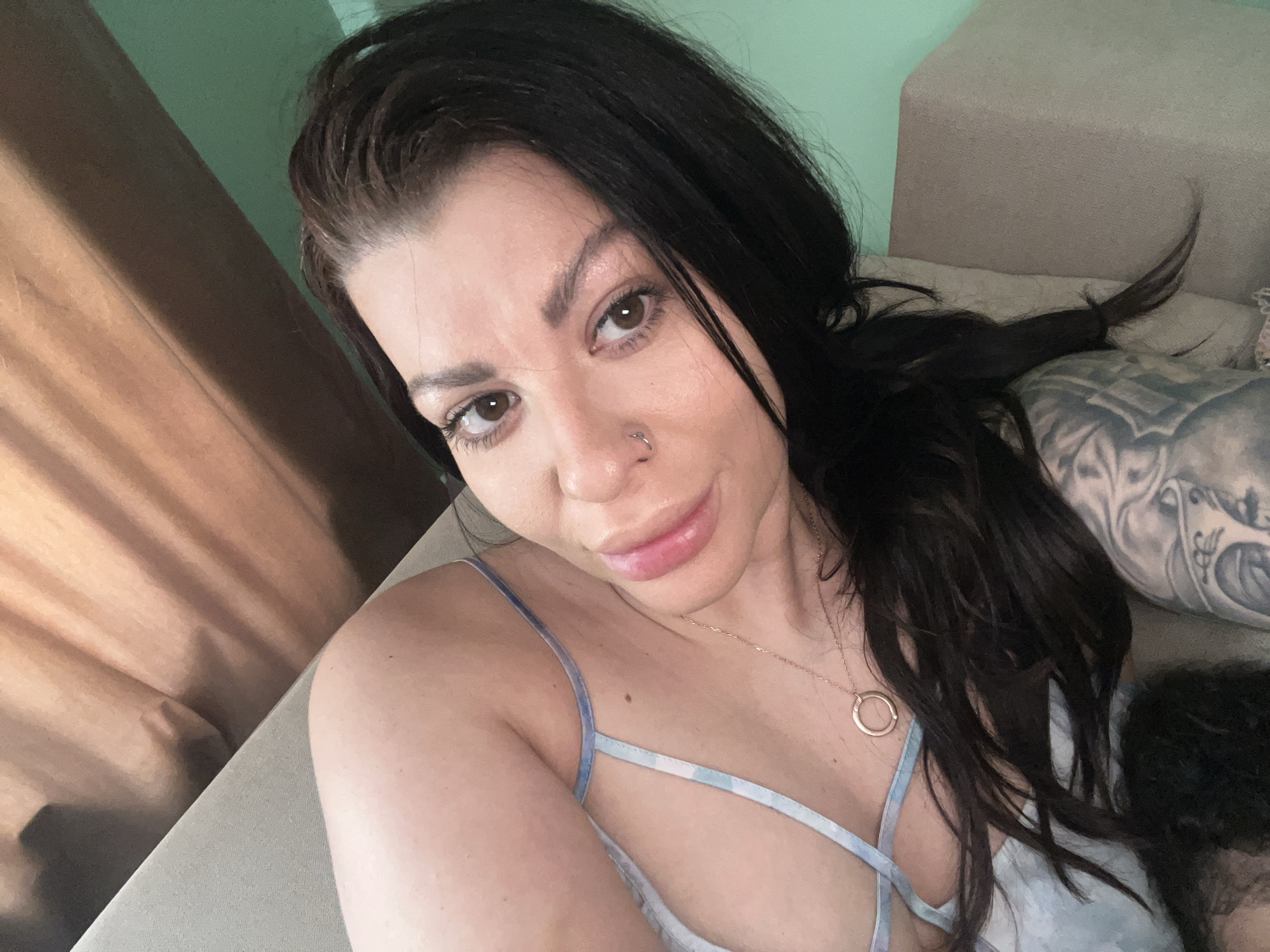 A woman with long, dark hair, brown eyes, and a nose ring is taking a selfie. She is wearing a light blue and white patterned slip dress and a gold necklace with a circular pendant. A tattoo is visible on her arm, and she appears to be lying down on a couch or bed. The background is a light green wall and a textured wooden surface. The scene is indoors, likely in a residential setting, and provides no specific location in Plovdiv, Bulgaria.