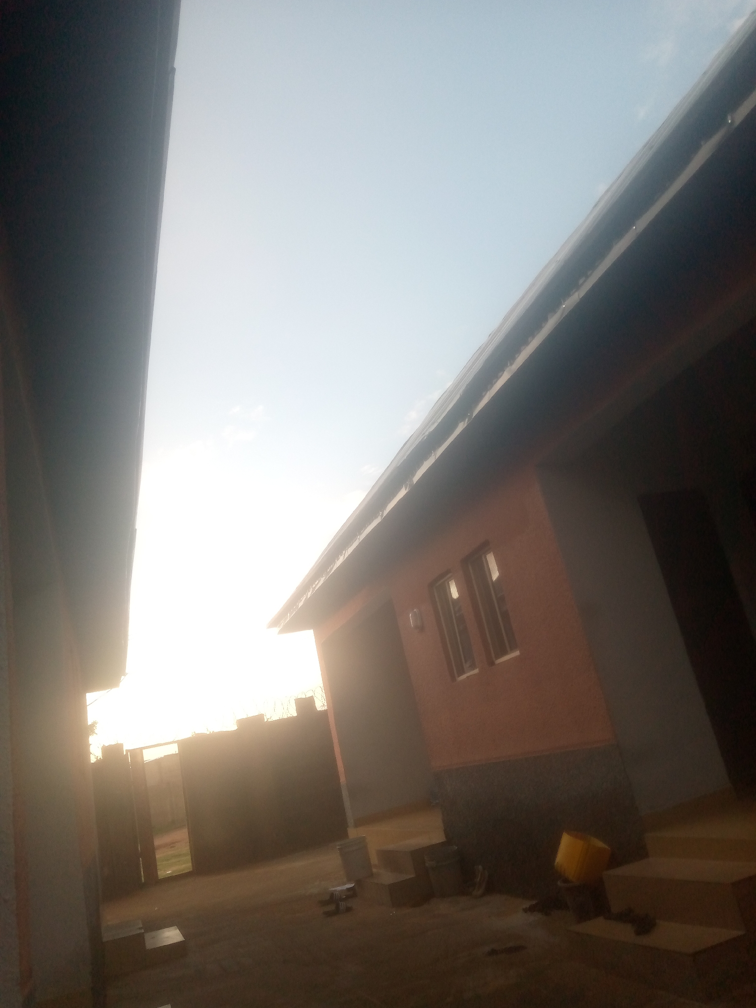 The image depicts an outdoor scene, likely within a residential or commercial compound, taken with a tilted perspective. The primary light source, presumably the sun, is positioned to the far left, creating strong backlighting that results in significant lens flare and deep shadows, particularly on the left side of the frame. The sky transitions from an overexposed white glare near the light source to a clear, pale blue with faint cloud wisps towards the upper right.
On the left, a large, dark vertical structure, possibly a wall or side of a building, occupies the left quarter of the image, mostly in deep shadow. To the right, a building with a salmon-pink or reddish-brown upper exterior wall and a dark grey or black lower foundation or wainscot is prominent. This building features two rectangular windows with white frames that reflect the bright sky. A dark roof, exhibiting minor indentations or features along its edge suggestive of a gutter system, overhangs the wall.
In the foreground and mid-ground, the ground is unpaved, appearing as compacted earth or gravel. Several dark, cubic objects, resembling wooden steps or stacked boxes, are arranged near the base of the building on the right. A bright yellow plastic bucket and additional dark, unidentifiable objects are scattered on the ground.
In the background, a tall, dark perimeter wall or fence, topped with what appears to be barbed or razor wire, spans across the scene. An open gateway or entrance is visible within this wall, leading to a brighter, unpaved area beyond. No people are present in the scene. The overall impression is that of a functional, possibly developing or utilitarian outdoor space, likely experiencing sunrise or sunset.