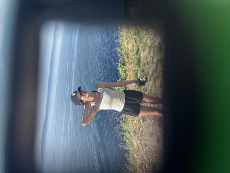 A young female is observed through an optical device's view, framed by dark, soft-edged borders. The scene is set on a grassy cliffside in Palos Verdes Estates, United States, overlooking the Pacific Ocean.
The female, appearing as a young adult, stands with her body angled slightly to the right, her head turned to gaze leftward across the ocean horizon. She wears a dark baseball cap with a light-colored logo, a light grey tank top, and black athletic shorts. Her right arm is raised with the hand positioned behind her neck, while her left hand holds a dark object with green accents, possibly a water bottle or small container.
The immediate foreground consists of dry, sparse grass and light green vegetation on a sloping cliff. A wide, calm blue ocean dominates the midground, its surface showing subtle ripples. Hazy mountain ranges or distant landmasses are visible on the far horizon under a clear, light blue sky. The scene is illuminated by bright daylight.