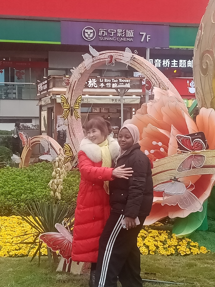 Two women pose for a photo in front of a decorative display. The woman on the left wears a red coat with a fur collar and a yellow scarf, while the woman on the right wears a black jacket and pants. The display features a floral design with butterflies and an arched structure adorned with lights. Behind them is a building with signs that read "SUNING CINEMA" and "LI RUO TAO YOGURT." Yellow flowers are planted in front of the display.