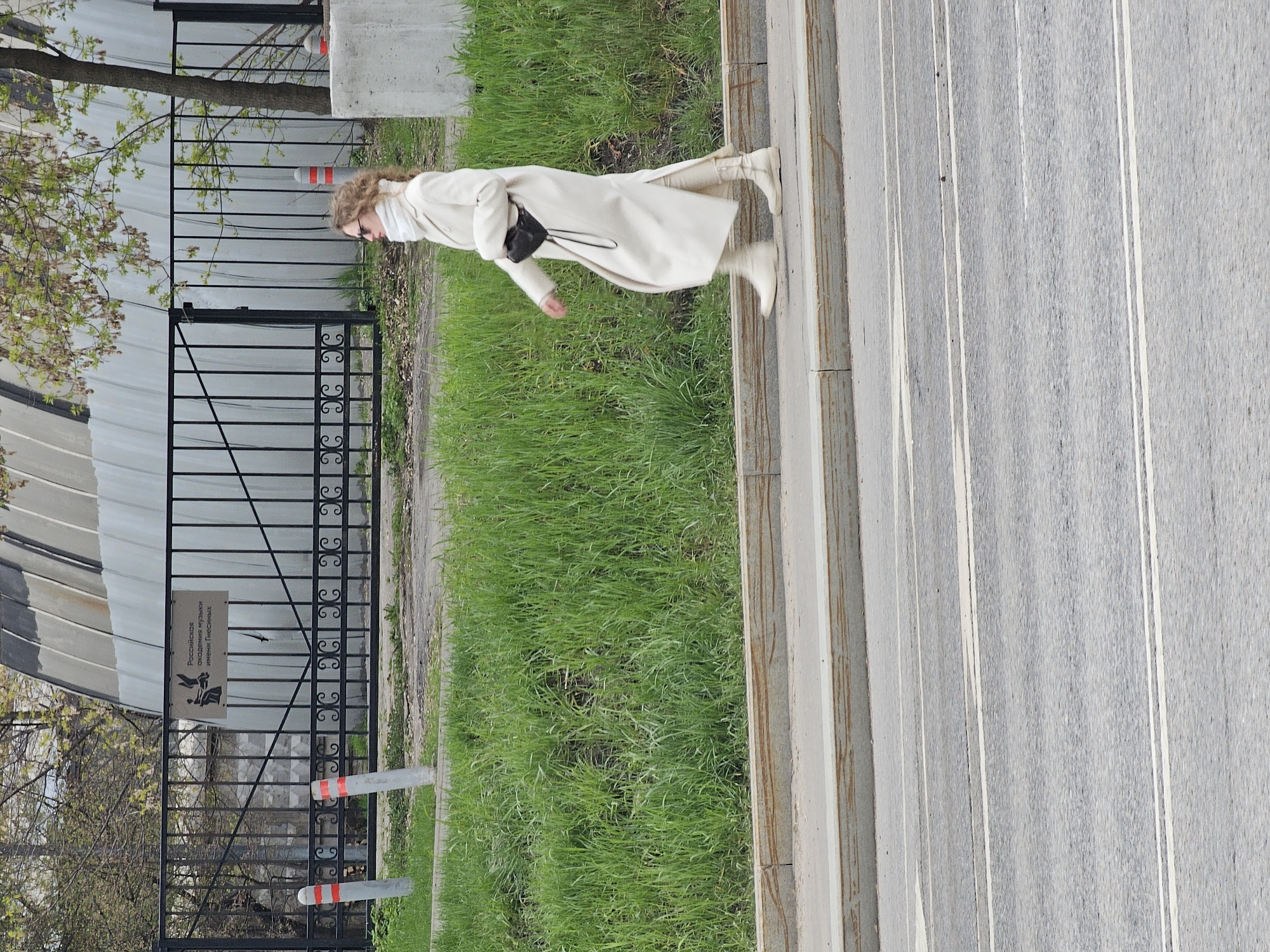 The image captures a woman walking outdoors in Moscow, depicted in profile and moving from right to left across the frame. She is dressed in a long, cream-colored or off-white coat, light-colored boots, and glasses, with curly light brown hair. She carries a small black cross-body bag.
She walks on a light-colored, possibly concrete or paved, pathway. Adjacent to the path is a strip of vibrant green grass. Further into the background, a black metal fence with decorative scrollwork runs horizontally. Behind the fence, a building or structure with a light-colored, possibly corrugated metal, facade is visible. A tree with fresh green spring leaves stands behind the fence. Two red and white striped bollards are positioned near the fence.
A rectangular sign is attached to the black metal fence. The visible Cyrillic text on the sign reads: "Российская академия музыки имени Гнесиных," which translates to "Russian Academy of Music named after Gnessin." This detail provides a strong location cue within Moscow.
The scene takes place during the daytime, under what appears to be overcast conditions, given the soft, diffused lighting. The overall atmosphere suggests a casual moment in an urban or institutional setting.