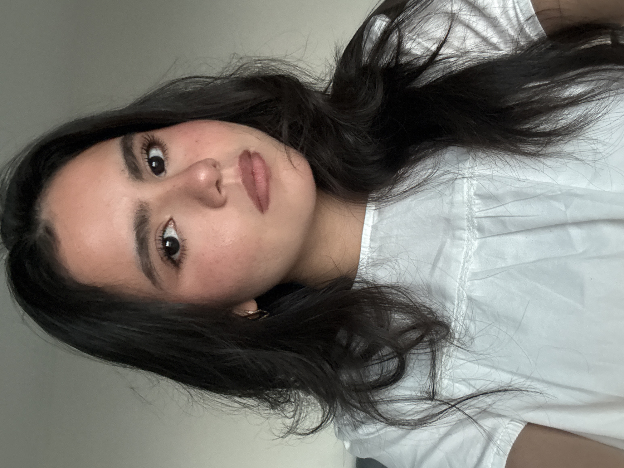 A young woman with dark hair and fair skin is captured in a close-up, head-and-shoulders portrait. She is wearing a white top with ruffled detailing around the neckline and sleeves. The background is a neutral, light gray wall, suggesting an indoor setting. Her makeup includes a touch of blush on her cheeks and lipstick on her lips. She has dark, prominent eyebrows and appears to be looking directly at the camera. A small gold hoop earring is visible in her left ear. The lighting is soft, casting gentle shadows on her face. There is no discernible information about the location within the city of Austin, United States of America.