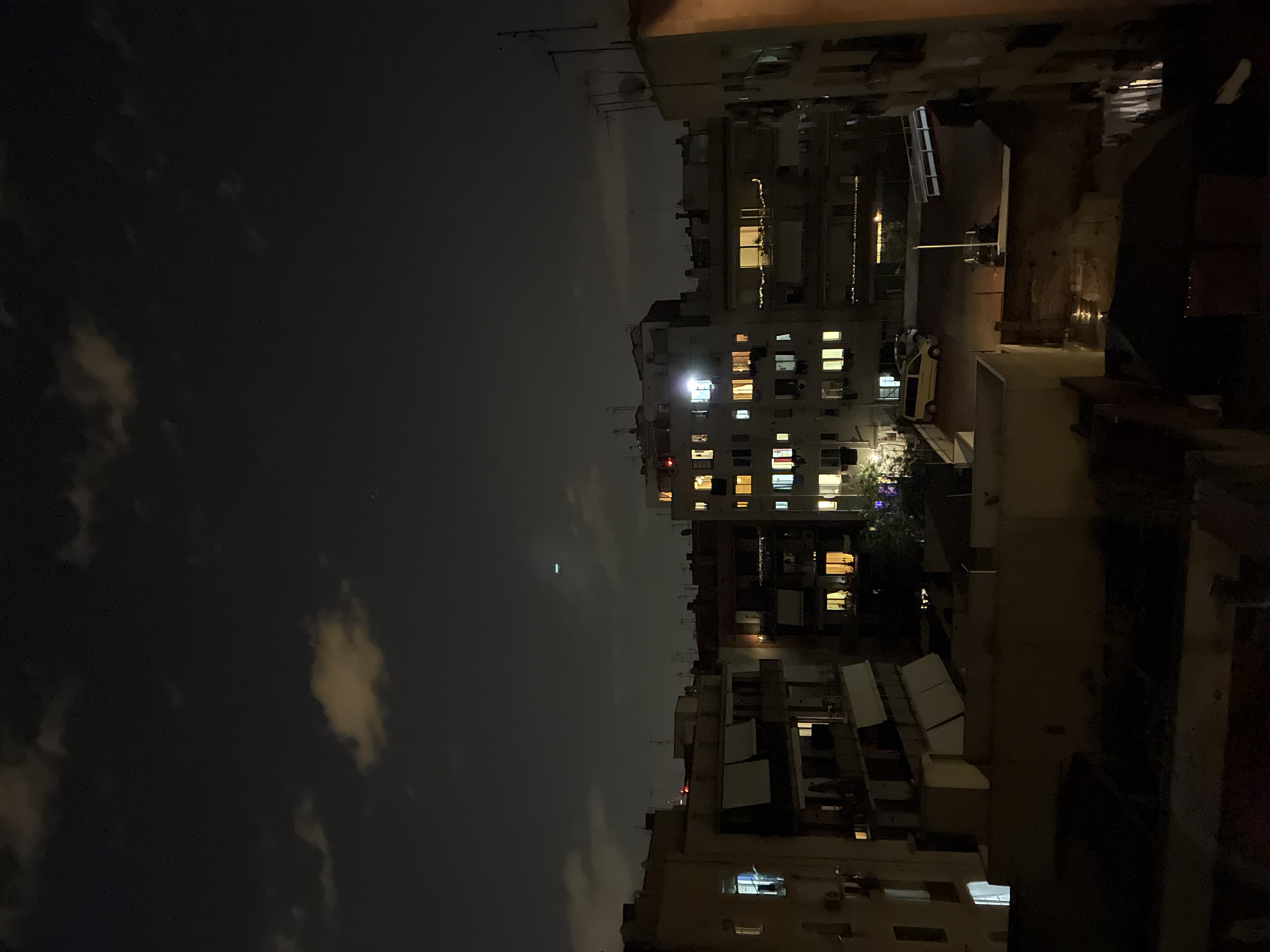 The image displays an urban night scene in Barcelona, Spain, viewed from an elevated perspective, likely a balcony or rooftop, looking across multiple residential or mixed-use buildings. The sky is dark, partially covered by scattered clouds. Numerous windows across several multi-story buildings are illuminated with warm yellow and white lights, indicating occupancy.
Notable details include a distinct elongated green light in the sky, possibly from an aircraft or a distant fixture, and a tiny bright dot further up. Between the buildings, a yellow vehicle, possibly a taxi, is visible on what appears to be a street or alley. Many rooftops feature satellite dishes and air conditioning units. Some visible ground surfaces and building elements appear wet, suggesting recent rain. Patches of dark foliage or trees are subtly illuminated by ambient light. No people or discernible text are visible, depicting a quiet, characteristic European city night.