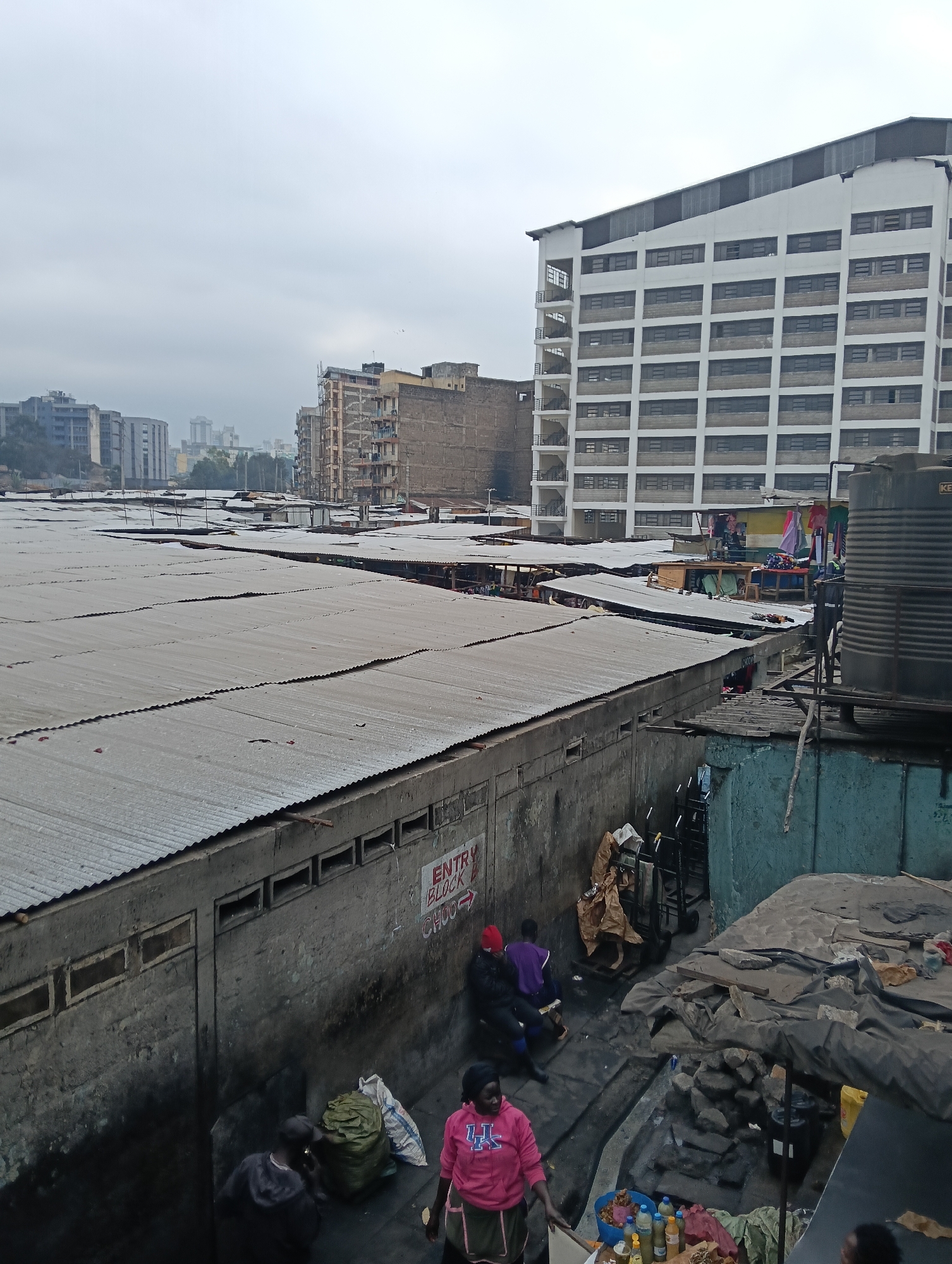 This image depicts a bustling market area in Nairobi, Kenya, likely in a densely populated or informal settlement. In the foreground, a rough, uneven ground surface is visible, with scattered debris and a large blue plastic basin filled with bottles of various liquids, possibly food or drinks. Several individuals are present in this area, with one person wearing a pink hooded sweatshirt and a green apron, appearing to be engaged in some activity. To the left, two people are seen near large bags of what appears to be produce or goods.
The middle ground reveals rows of corrugated metal roofs covering market stalls. These roofs are irregular and appear aged, with some gaps and imperfections. Adjacent to the market stalls is a concrete structure with rectangular openings, likely ventilation or small windows. A sign on this wall reads "ENTRY BLOCK CHOO".
In the background, several multi-story buildings are visible, indicating an urban setting. One prominent, large white building with many windows dominates the right side of the frame. Clothes hang from lines on smaller structures behind the market stalls, adding to the sense of active commerce and daily life. A large dark water tank is situated on a platform to the right of the white building. The overall environment suggests a crowded, active, and somewhat improvised market scene within an urban context. The sky is overcast, suggesting a dull or cloudy day.