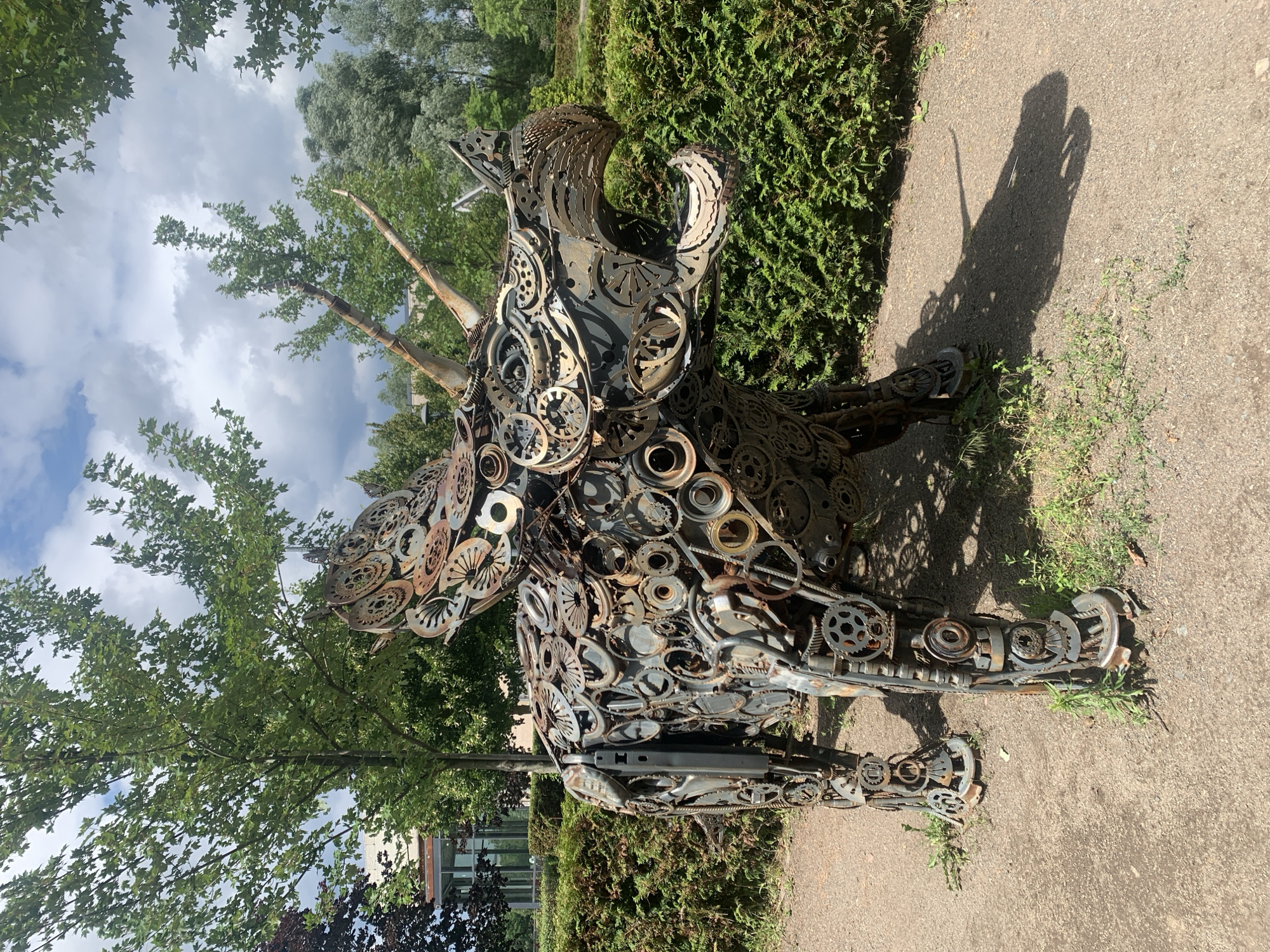 A metal sculpture resembling a dinosaur, constructed from gears and other recycled machine parts, stands outdoors on a gravel path in Bucha, Ukraine. The sculpture is positioned in front of a neatly trimmed green hedge. Trees with green foliage and a cloudy sky are visible in the background. The sculpture casts a shadow on the path.