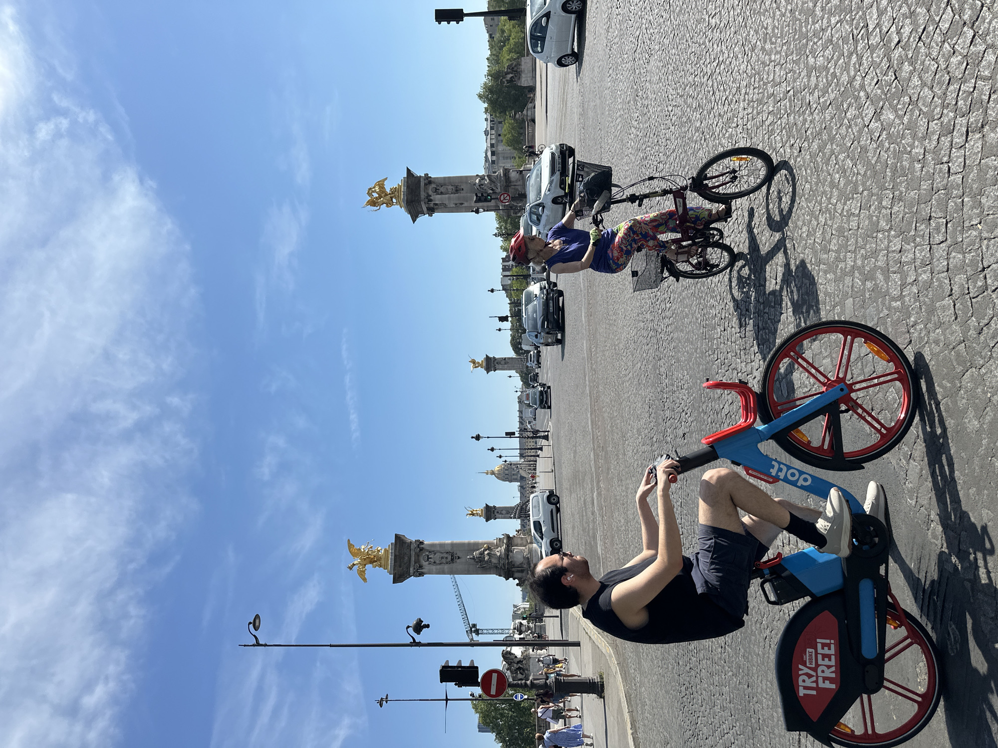 This media file shows a sunny day in Paris, France. The main subjects are two people on bicycles. In the foreground, a man wearing a black tank top and dark shorts is sitting on a blue and red Dott electric bicycle. He is looking to his right and holding onto the handlebars. The bicycle has a "TRY FREE!" sticker on the front. Behind him, a woman with blonde hair, wearing a purple top, colorful leggings, and a red helmet, is riding a red bicycle. She is looking to her left.
The scene is a cobblestone street with other Dott electric bicycles parked nearby. In the background, iconic Parisian landmarks are visible, including the ornate statues and golden figures adorning the Pont Alexandre III bridge. Cars and people are also present, suggesting a bustling urban environment. The sky is a clear blue with a few wispy clouds. The lighting indicates it is daytime, likely late morning or early afternoon, given the bright sunshine and distinct shadows. The overall mood appears relaxed and active, capturing a moment of leisure and exploration in the city.