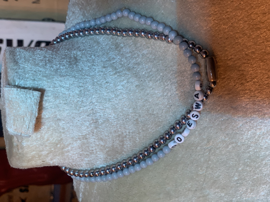 A close-up image of a silver and light blue beaded necklace. The necklace is made of two strands of beads, one silver and one light blue. The silver beads are round and metallic, while the light blue beads are slightly oval-shaped and have a matte finish. The beads are strung together with a silver metal thread. The necklace has a silver clasp and is lying on a light gray, fuzzy surface. The necklace spells out the word "SWAG" using white letter beads. The image was taken indoors, likely in a home or shop. The coordinates provided (latitude: 7.282, longitude: 5.161) are in the city of Ita Oniyan, Nigeria. There are no people or actions in the image.