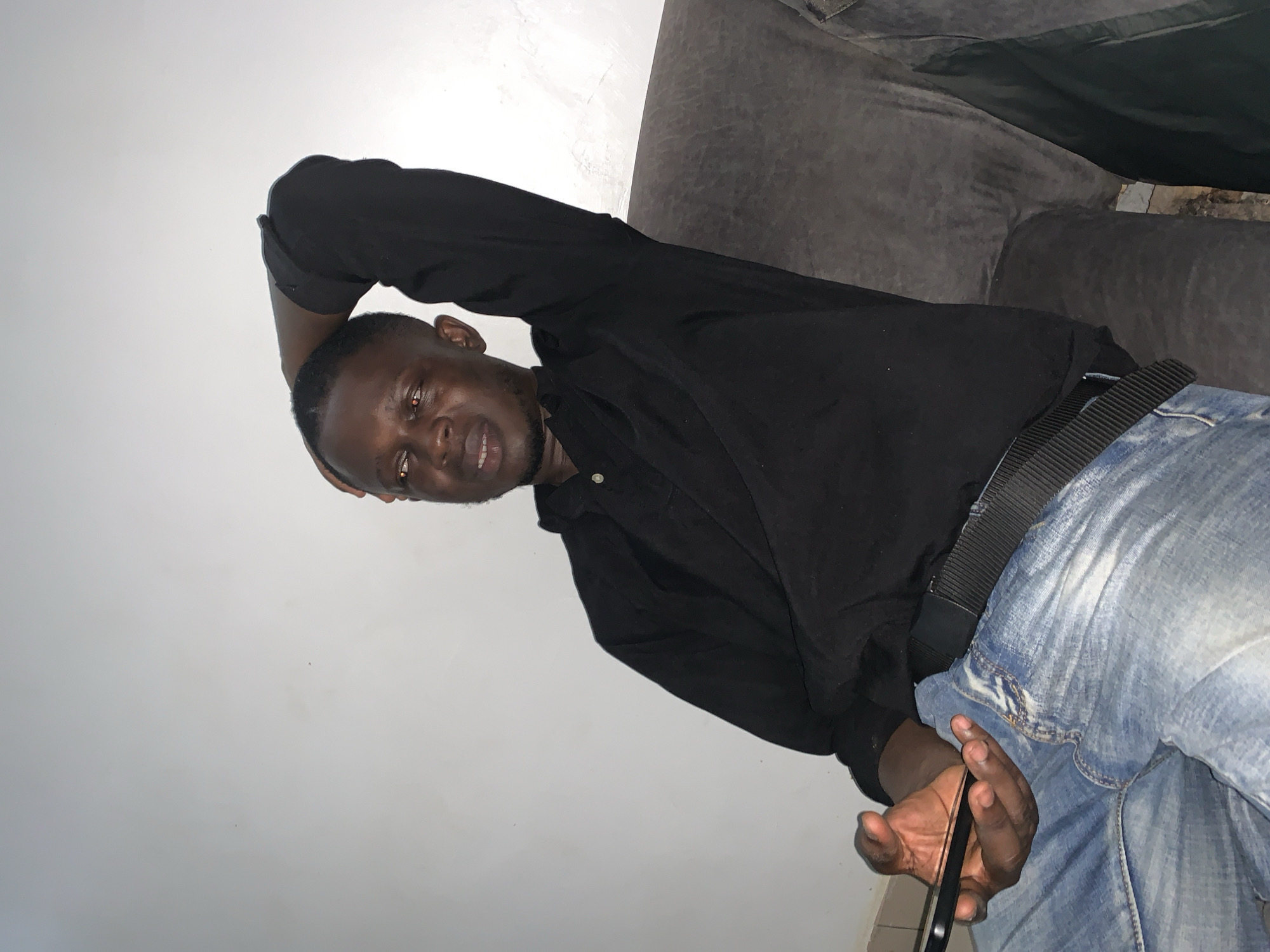 A dark-skinned man with short black hair is seated on a gray sofa in front of a white wall. He is wearing a black collared shirt and light blue distressed jeans with a black belt. His left arm is bent with his hand resting on the back of his head, and his right hand is holding a black smartphone. He is looking towards the right with his mouth slightly open. The background is a plain white wall, and the lighting suggests an indoor setting. This scene is consistent with a casual living room environment, potentially in Abuja, Nigeria.