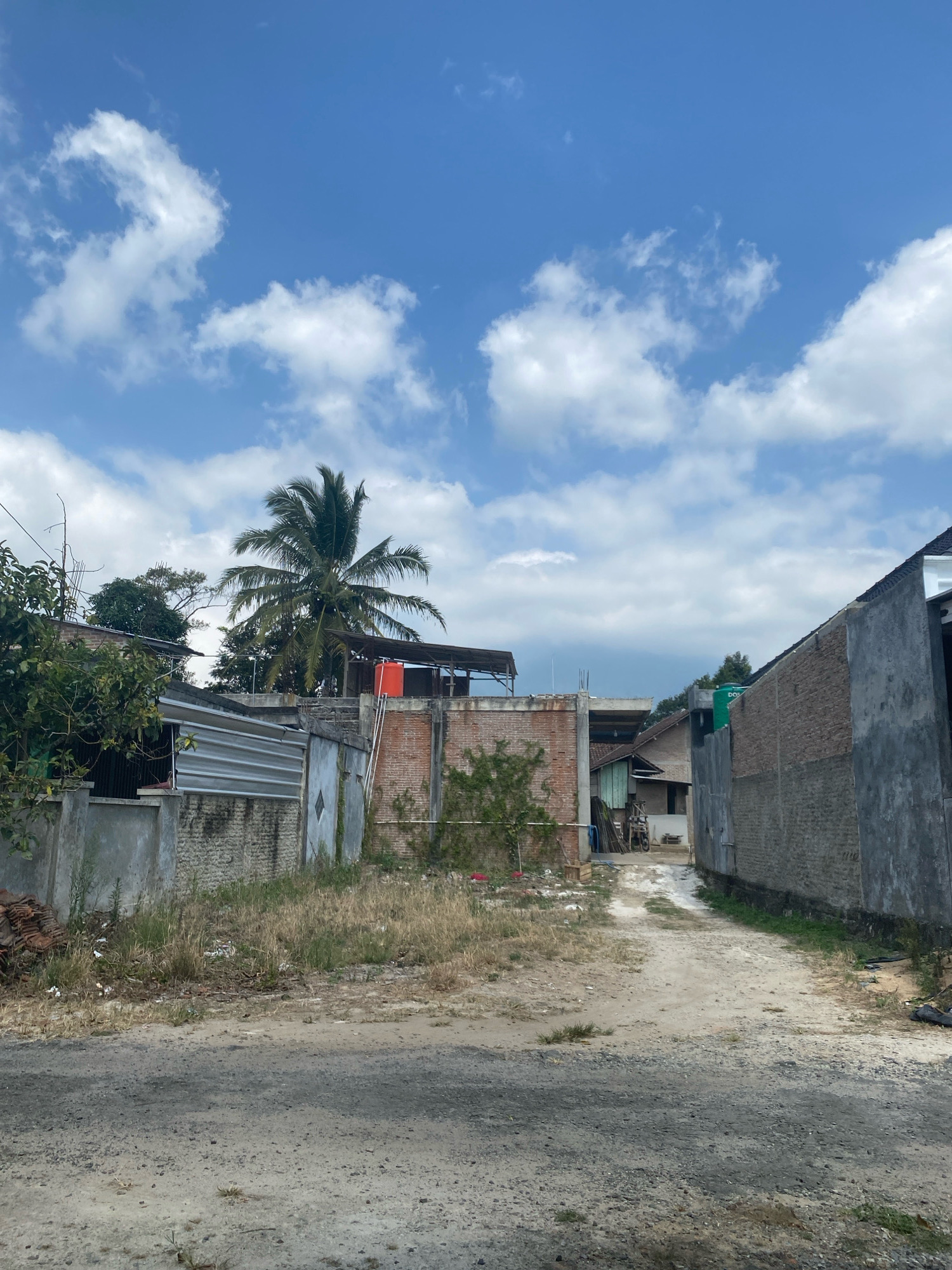The image depicts a dirt road in a semi-urban area of Kota Dalam, Indonesia, under a bright blue sky with scattered white clouds. On the left side of the road, there is a low concrete wall topped with a corrugated metal fence and rough stone blocks. Behind this, a brick building with a weathered facade is visible, partially obscured by lush green foliage and a mature palm tree with a bright orange water tank mounted on its roof. Further back, there are more buildings constructed from brick and concrete, with some exhibiting exposed construction materials. A wooden structure with a sloped roof stands adjacent to a brick wall.
To the right of the road, a tall, rough concrete wall dominates the foreground, its texture contrasting with the smooth, dark grey asphalt of the road. Beyond this wall, a brick building with a darker grey concrete facade is visible. Further down the road, more buildings can be seen, including a structure with a dark grey pitched roof and a partial view of a green water tank. The road itself is unpaved, with patches of dry grass and sparse vegetation growing along its edges and in the surrounding undeveloped land. Debris, including scattered litter and piles of broken roof tiles, is present in the foreground and along the roadside. The scene appears to be a developing or partially completed construction area.