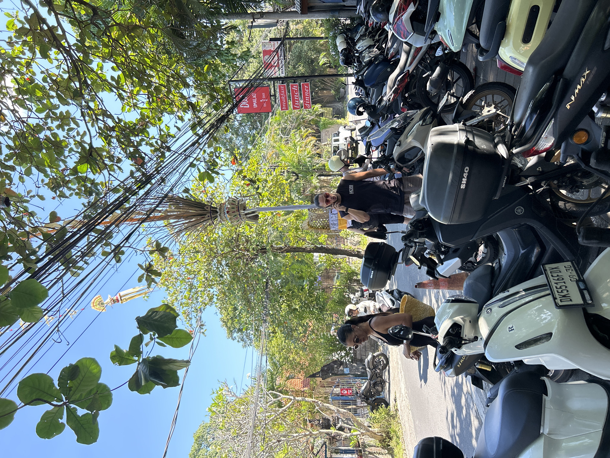 A street scene in North Kuta, Indonesia, captures a bright, sunny day with a clear blue sky. The foreground is dominated by a row of parked motorcycles, predominantly scooters, with some bikes also visible further down the street. A woman wearing a black tank top and sunglasses, carrying a woven straw bag, is walking on the sidewalk on the left side of the image. To her right, a man wearing a black tank top with the word "DEUS" visible, and sunglasses, is on his phone. Behind him, a white SUV is parked on the street.
Overhead, a dense network of electrical wires crisscrosses the frame, with lush green trees providing shade. Decorative Balinese umbrellas, known as "umbrellastands" or "pawitra", are suspended from some of the wires, indicating a festive or cultural event. A signpost with red rectangular signs advertising "breakfast", "lunch", and "dinner" is visible, suggesting the presence of a restaurant or food establishment. The architecture visible in the background, including a gate with intricate carvings, is characteristic of Balinese design. The overall impression is a vibrant streetscape in a tropical location, bustling with activity and local culture.