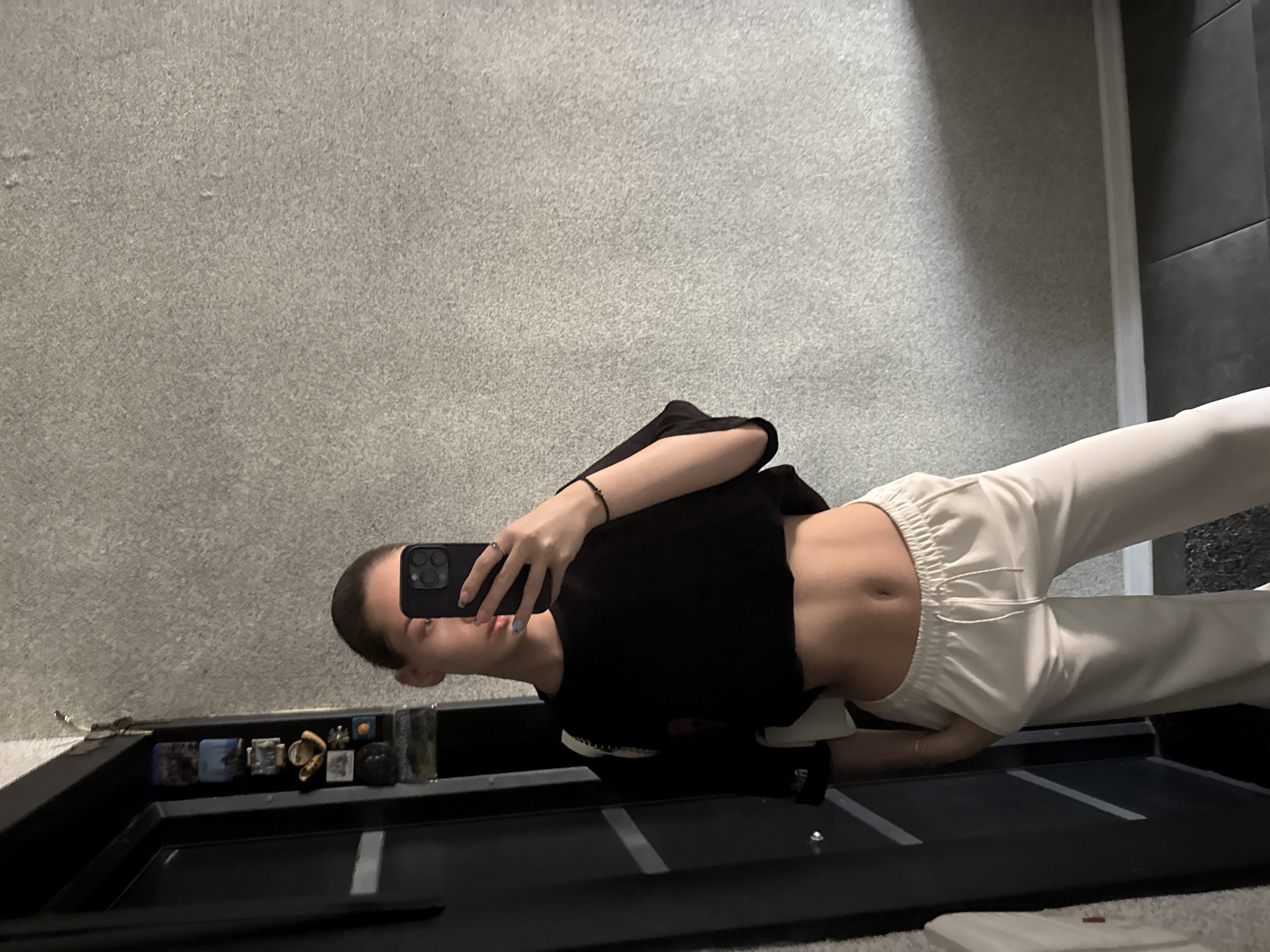 A woman is taking a selfie in front of a mirror. She is wearing a black crop top and white pants. The background consists of a gray wall and tiled area. There is no indication of location in Sofia, Bulgaria.