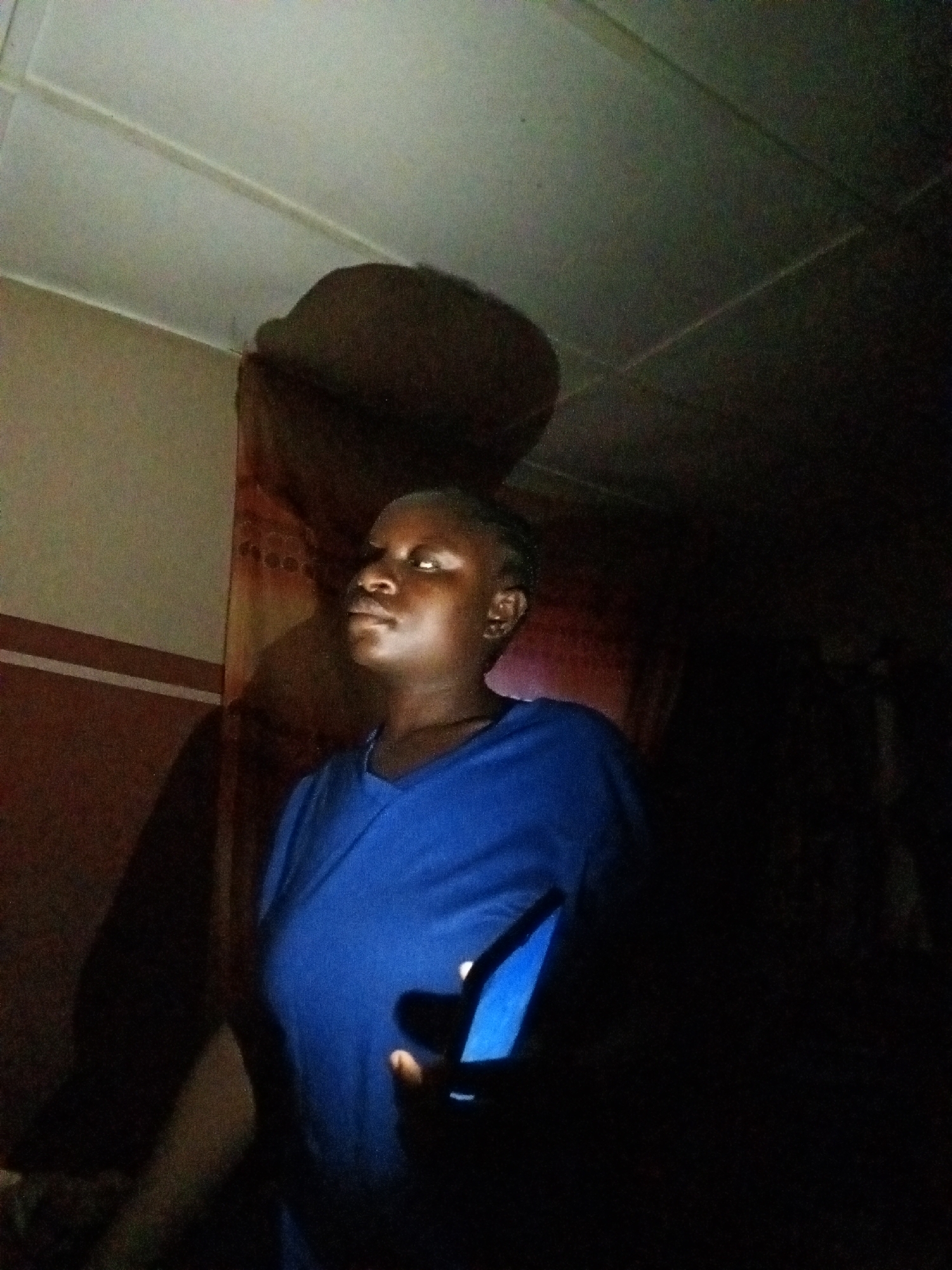 A young woman, likely of African descent, is captured in a dimly lit indoor setting, possibly a bedroom. She is positioned facing left, with her head turned slightly towards the camera. Her dark skin is illuminated by an artificial light source from her right, casting a noticeable shadow on the wall behind her. She is wearing a bright blue V-neck t-shirt. Her left hand is visible holding a mobile phone, which emits a blue light that reflects on her t-shirt and illuminates her hand. The background suggests a room with a white, paneled ceiling. A curtain or fabric hangs on the wall to her left. The overall impression is that of a candid, low-light portrait taken with a mobile phone.
