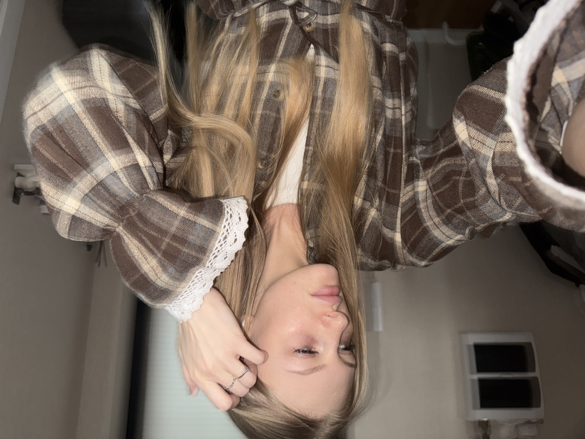 A young woman with blonde hair is pictured from an inverted angle. She is wearing a brown and beige plaid shirt with lace trim on the sleeves. Her right hand rests on her head. A ring is visible on her finger. The background includes a wall, a black object out of focus, and what appears to be a window unit. The city of Perm, Russia, is not discernible from the available details in the image.