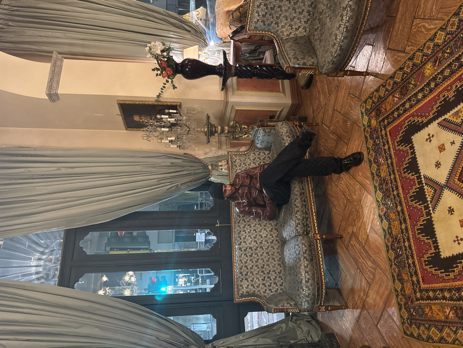 The image depicts the opulent interior of a historic building in central Milan, Italy, characterized by grand architectural features and large street-facing windows.
**People:**
One male individual is seated on a decorative sofa. He wears a dark beanie, a reddish-brown long-sleeved shirt with an abstract pattern, dark patterned trousers, and dark dress shoes. His legs are crossed at the ankles, and he is looking down, seemingly at an object held in his hands, which is not clearly identifiable.
**Subjects and Objects:**
The room features a polished light brown herringbone-patterned tile floor covered by a large Oriental rug with complex geometric designs in shades of red, beige, black, and yellow.
A prominent two-seater antique-style sofa, with a gilded, carved wooden frame and dark teal/gold patterned upholstery, occupies the center-left. The individual is seated upon it. To the right, a matching antique-style armchair is visible.
Two tall, dark-framed windows with arched transoms are draped with heavy, light blue/grey floor-length curtains. Through the windows, an urban street scene at dusk or night is visible, including city lights, reflections, and a distinct green traffic light.
The walls are painted a warm, light beige, adorned with classical architectural details such as moldings and pilasters. A framed portrait painting of an older male figure hangs on the wall.
Ornate decorative pieces include a large, non-hanging crystal chandelier-style lamp positioned on a tall, gilded pedestal that features a sculptural element, and a tall, dark, twisted pedestal holding a dark vase filled with white, red, and orange flowers and green foliage.
**Actions and Interactions:**
The male individual is in a state of repose, focused downwards, with no apparent interaction with other individuals or the immediate environment beyond his seated posture.
**Environment:**
The environment is one of classical luxury and elegance, suggestive of a high-end establishment such as a grand hotel lobby, a historical cafe, or a private residence in a prominent urban location. The artificial interior lighting creates a warm ambiance that contrasts with the cooler tones of the exterior street scene.