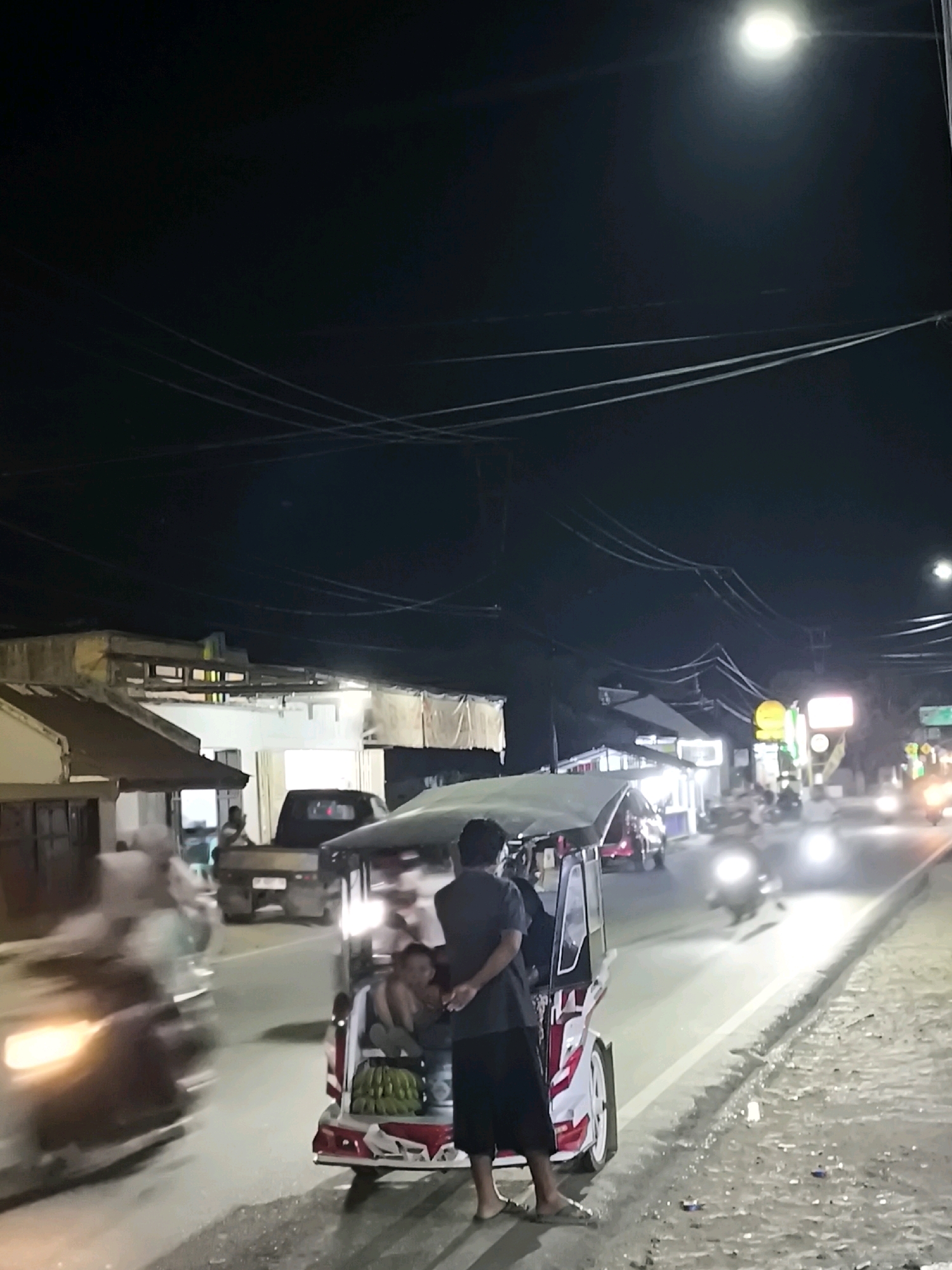 A nighttime, medium shot captures a street scene in Moluo, Indonesia, featuring a busy intersection with several vehicles and a pedestrian. In the foreground, a colorful, three-wheeled vehicle, possibly a bemo or an auto-rickshaw, is parked on the right side of the road. It is adorned with red and white decals and has a canopy. A bunch of bananas is visible in the rear compartment. Two individuals are seated inside the vehicle. A man, wearing a dark shirt and shorts, stands near the open side of the vehicle, facing away from the camera with his hands behind his back. To the left of the bemo, a blurred motorcycle speeds by, its headlights illuminating the road. Further down the street, a black pickup truck is parked. The background depicts a row of buildings with illuminated signs and awnings, suggesting a commercial area with shops or food stalls. Overhead, power lines crisscross the dark sky, with a bright street lamp providing illumination at the top of the frame. The overall impression is of a vibrant, active night market or street scene.