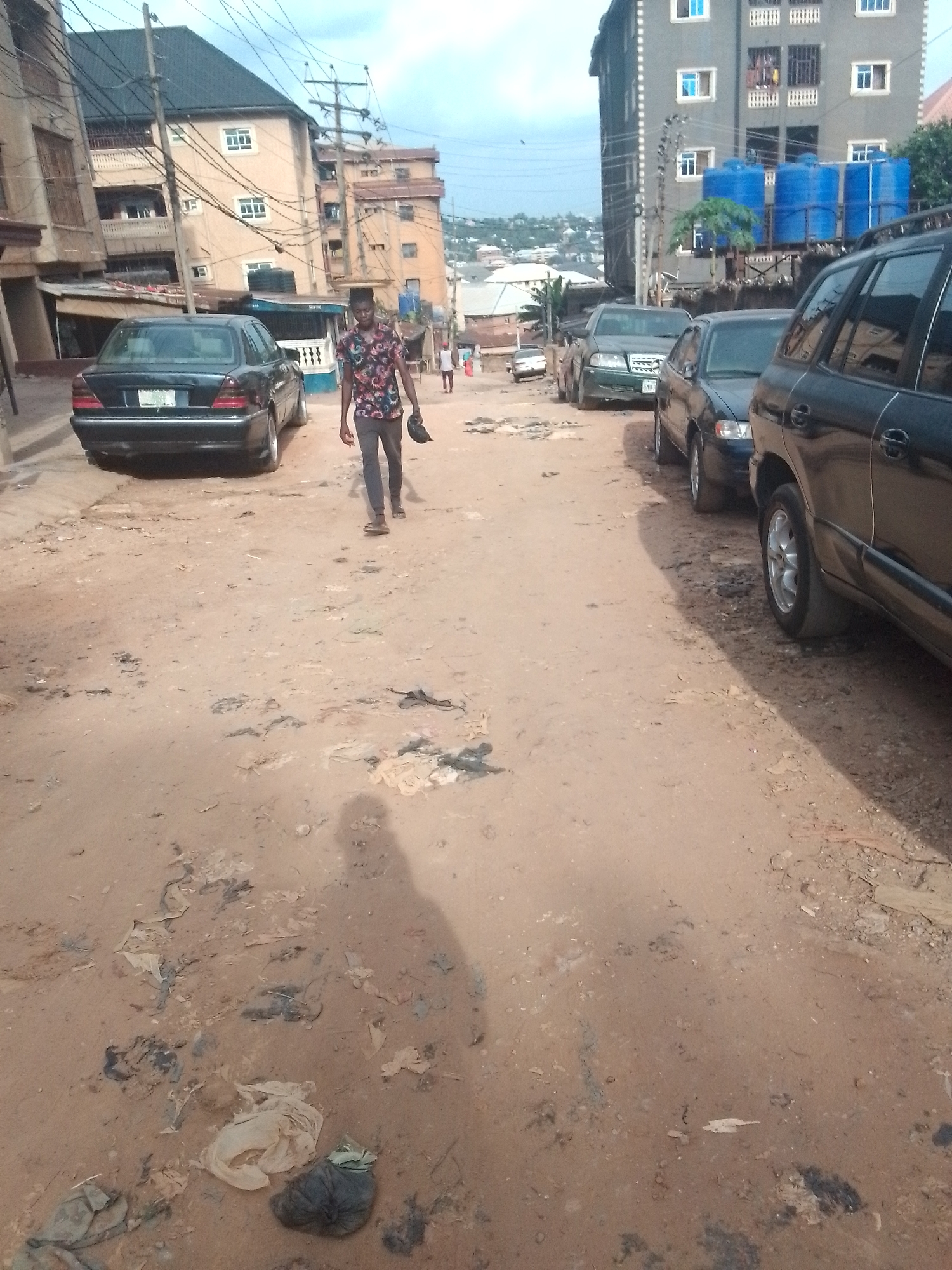 The scene is a dusty, unpaved street in Onitsha, Nigeria. Debris litters the ground. A man walks towards the camera, holding a dark object in his left hand. Cars are parked along the right side of the street, while one is parked on the left. A building with blue water tanks on the roof is visible in the background. Other buildings line the street, some taller than others. Power lines crisscross overhead.