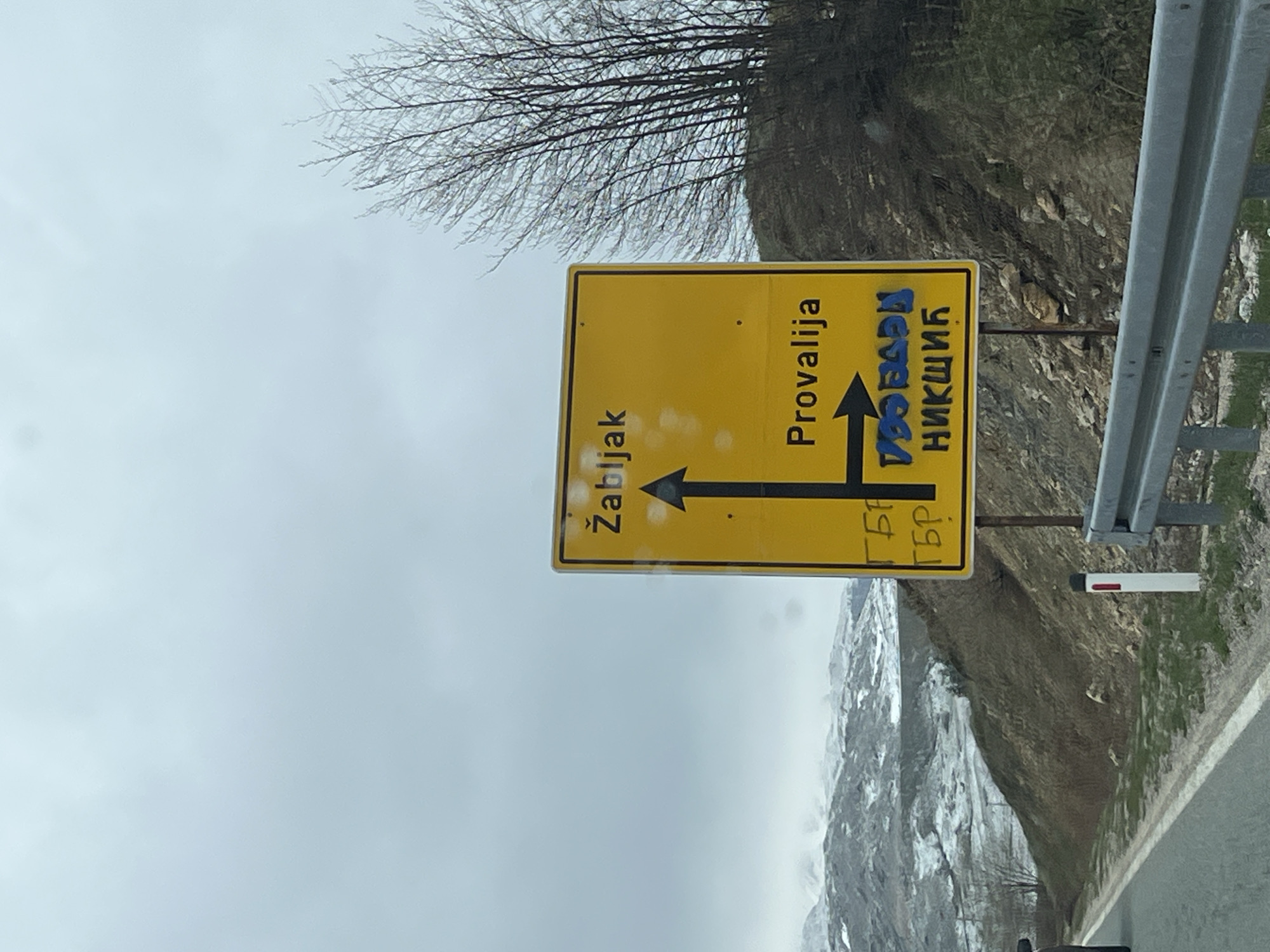 The image captures a roadside scene, likely from a vehicle, featuring a yellow directional sign in a mountainous region of Montenegro. The sign, rotated on its side, points towards several locations. An arrow pointing left indicates "Žabljak". An arrow pointing straight ahead indicates "Provalija". An arrow pointing right indicates "НИКШИЋ" (Nikšić), which has blue graffiti "ТПО БРАЋА" (TPO Braća) partially obscuring it. Faintly written at the bottom right of the sign, also in graffiti, are the letters "ГБР" repeated twice.
The setting is outdoors during an overcast day, suggested by the muted light and a grey sky filling much of the background. To the right of the sign, a grey metal road barrier or guardrail runs parallel to what appears to be a road. Beyond the barrier and the sign, the terrain is rugged and brown, leading up to a distant mountain range whose peaks are partially covered with snow. A bare-branched tree, indicative of winter or early spring, is visible in the upper left of the frame. The overall impression is a cold, remote, and natural landscape. The presence of the road sign explicitly linking to "Provalija" and "Nikšić" confirms the location context of Montenegro.