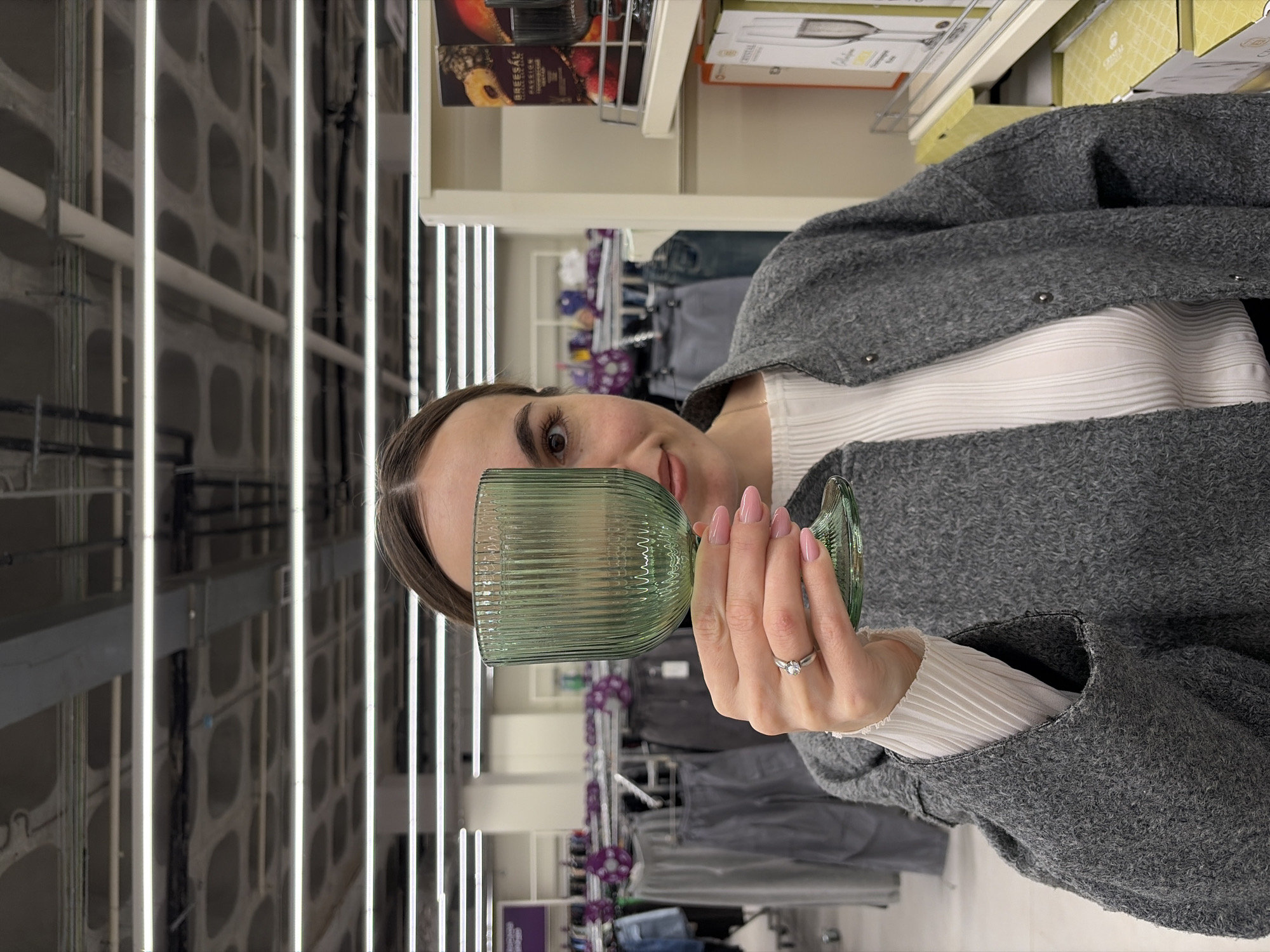 A woman stands in a store, holding a green glass in front of her eye. She is wearing a gray coat over a white shirt. Behind her, shelves of merchandise are visible. The store appears to be a department store or a large retail space, indicated by the rows of clothing and other goods. The background includes linear lighting fixtures typical of a retail environment. The specific location in Izhevsk, Russia, cannot be definitively determined from the visual information provided.