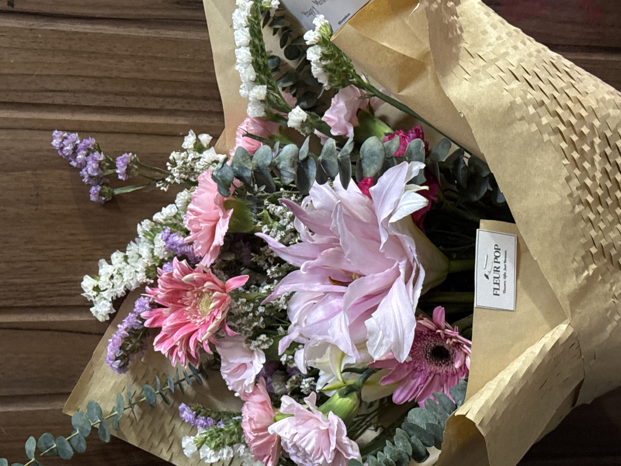 A floral bouquet in a kraft paper wrapper sits on a wooden surface. The bouquet contains pink gerbera daisies, pink carnations, white and purple statice flowers, eucalyptus leaves, and what appears to be a light pink lily. The kraft paper wrapper features a label with the text "FLEUR POP" and a textured honeycomb pattern on a portion of the packaging. The scene likely occurred in Davao City, Philippines as Fleur Pop is a local business located in the city.