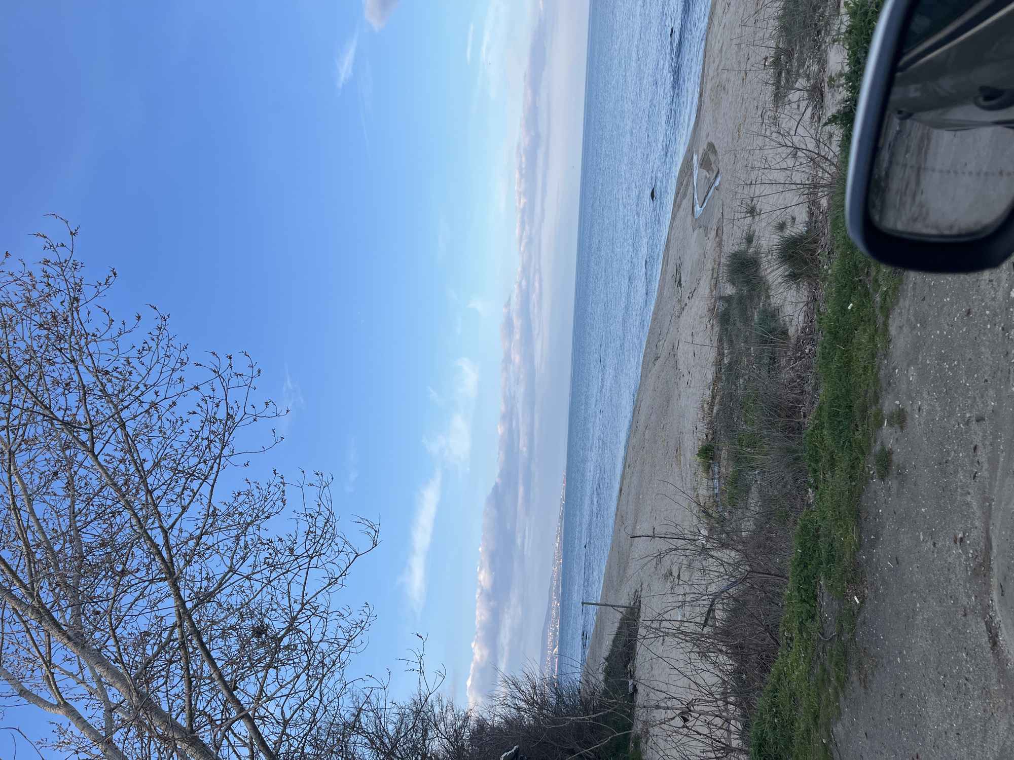 The image shows a seaside view in Varna, Bulgaria. The scene is captured from a vehicle, evidenced by the presence of a side mirror in the upper right corner of the image. The sea, under a partly cloudy blue sky, stretches along the horizon. A sandy beach is adjacent to the sea, with sparse vegetation growing along its edge. A paved road runs parallel to the beach. Bare trees are visible in the lower left corner of the image. In the distance, the city lights indicate a cityscape.