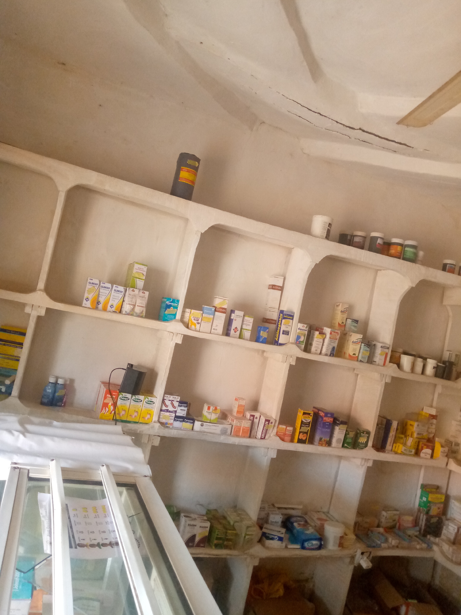 The image depicts the interior of a small pharmacy or drug store located in Kauru West, Nigeria. The pharmacy appears to be sparsely stocked, with shelves made of what looks like concrete or plaster, forming cubby-like compartments. These shelves are filled with various boxes and containers of medicines and pharmaceutical products. Some of the visible items include boxes of "CLINCEF," "Polygy" and "Pyrazinamide." There are also bottles of what appear to be liquid medication on a lower shelf. In the foreground, a white sheet or cloth covers a counter or display area, and part of a glass display case is visible. The ceiling shows signs of wear, with some cracks and staining. A cylindrical object with orange and yellow labeling is present on a high shelf. The lighting is dim, suggesting it might be daytime with limited natural light, or the artificial lighting is not very bright. The overall impression is of a functional but basic retail establishment in a rural or semi-urban Nigerian setting.