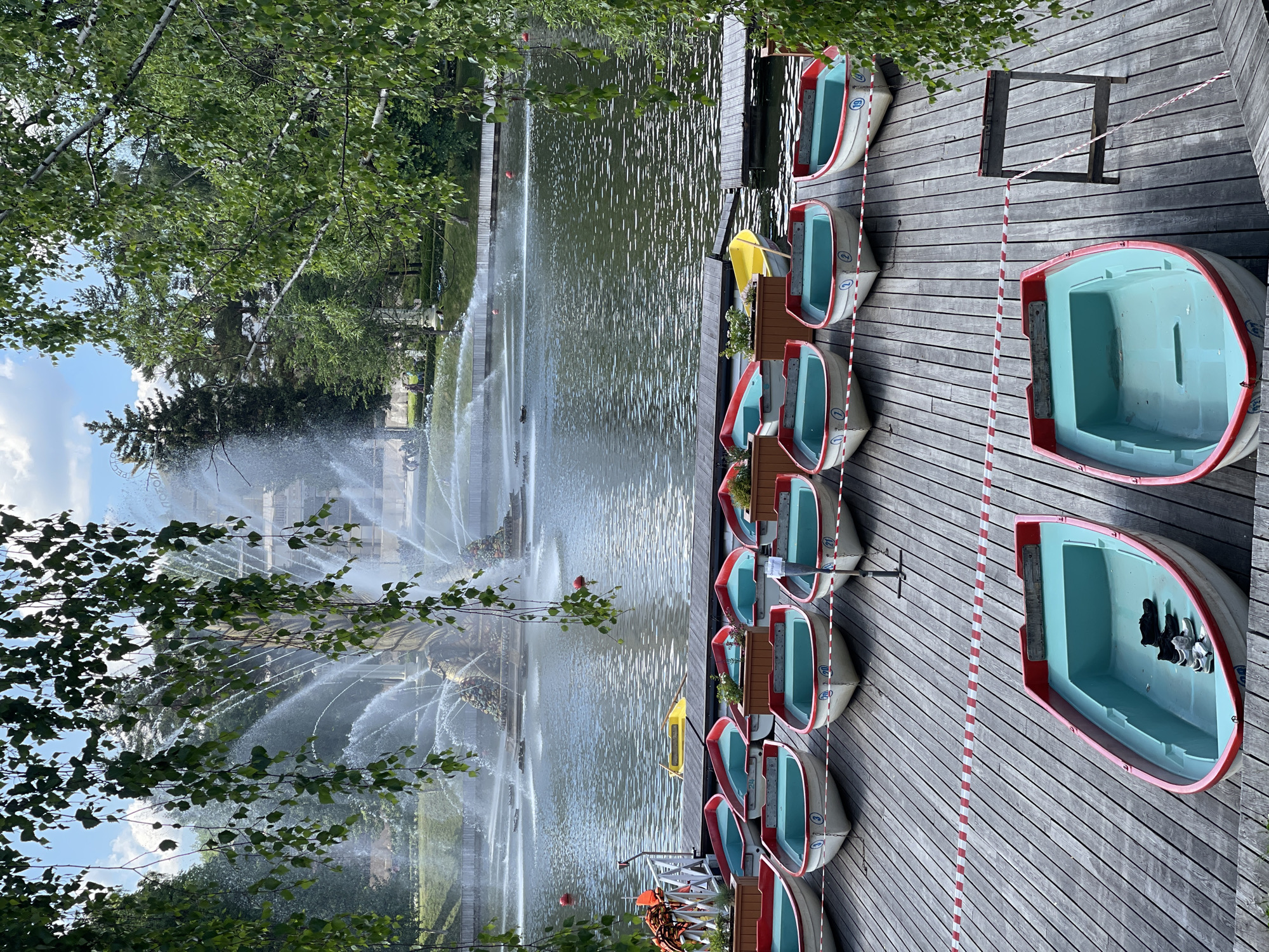 The image, rotated 90 degrees clockwise for proper viewing, displays an outdoor scene at a recreational body of water, likely a park or leisure lake. On the right, a wooden dock or deck is lined with numerous small, identical boats. These are white with red trim and light blue interiors, appearing to be rental pedal boats or rowboats. Some boats have numbers visible in blue circles, such as "10" and "2". A red and white striped tape runs along the dock, possibly marking a boundary or safety line. One boat on the dock contains a pair of shoes or sandals. A small yellow boat is partially visible near the dock.
On the left, a large, ornate fountain is actively spraying water high into the air and across the water's surface, creating multiple arcs and a misty atmosphere. Through the water spray, lush green trees and a bright blue sky with scattered white clouds are visible, indicating a clear, pleasant day. Faintly discernible in the background through the fountain's mist, the word "VICTORIA" can be made out, suggesting the name of the fountain or a nearby landmark. The scene evokes a sense of leisure and outdoor activity during daytime.
