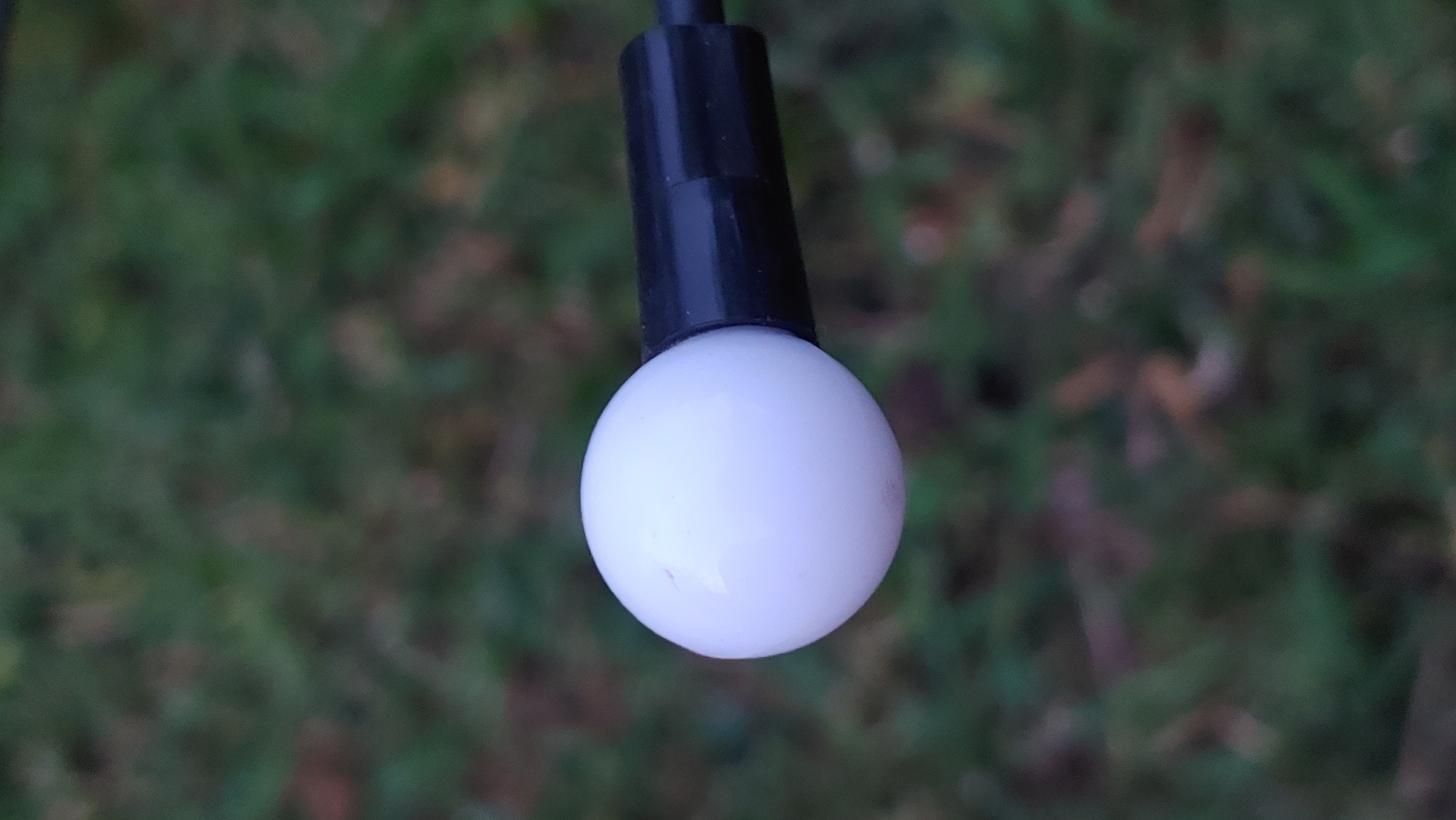 This media file features a close-up shot of a single white, spherical LED light bulb, suspended by a black connector against a blurred background of green grass. The bulb is the central focus, appearing smooth and matte, with a subtle sheen indicating light reflection. A small, faint reddish mark is visible on its surface. The black connector, cylindrical in shape, attaches to the top of the bulb and extends upwards, presumably to a wire or power source, though these are largely out of frame. The background is an out-of-focus expanse of green, suggesting an outdoor setting, possibly a lawn or garden. The lighting appears to be ambient, possibly natural daylight, given the soft illumination on the bulb and the lack of harsh shadows. There is no visible text within the frame. The overall impression is one of simplicity and focus on the object itself, with the natural setting providing a subdued contrast. The scene is static, with no discernible activity taking place beyond the presentation of the object. The location context of Mukachevo, Ukraine, does not provide specific visual cues within the image itself, as the scene is generic in its elements.
