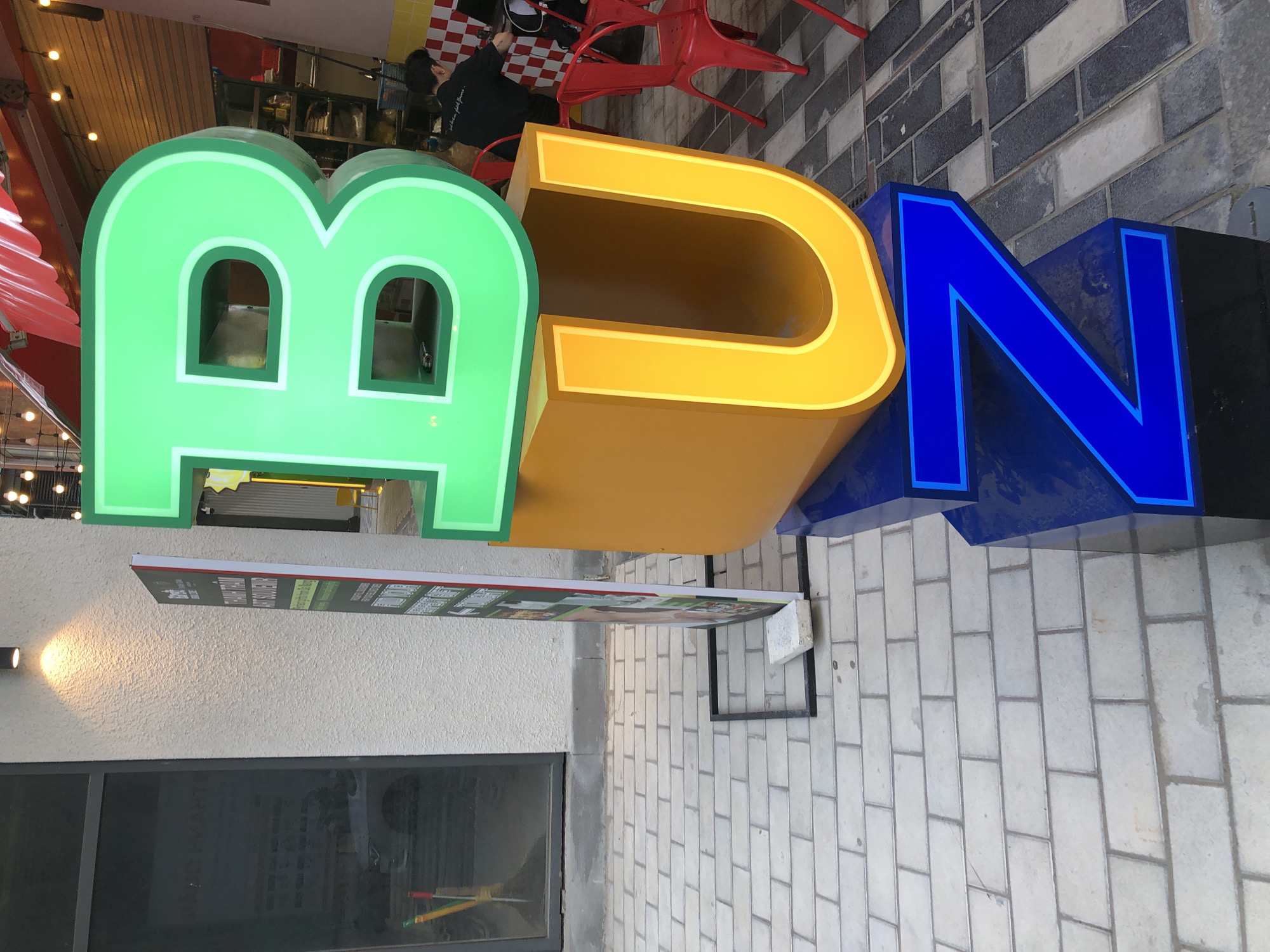 The image shows a close-up of a large, three-dimensional sign that spells "BUZZ" with illuminated letters. The "B" is bright green, the "U" is a glowing yellow, and the "Z" is a vibrant blue. The sign is positioned outdoors on a paved walkway made of light-colored bricks. In the background, there is a building with a textured wall and a dark-tinted window. Red chairs are visible to the right, suggesting an outdoor seating area. A person is partially visible in the background, seated at a table. A tall, thin sign with text and images stands to the left of the "BUZZ" sign. The overall impression is that of a commercial establishment, possibly a restaurant or cafe, in an urban setting.