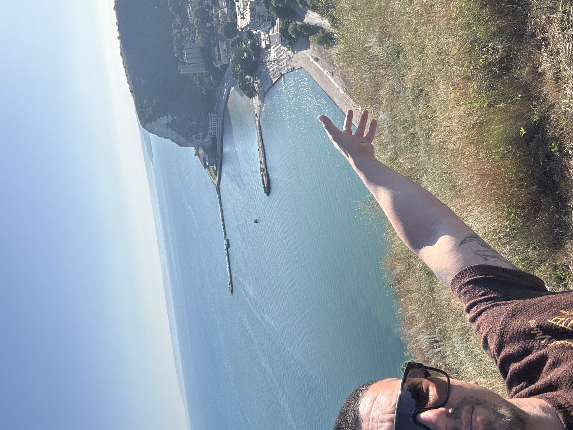 The media file is a selfie taken outdoors, with the photographer's arm extended and hand open. The scene is dominated by a vast expanse of calm, blue water, likely the sea, which reflects the bright sunlight. In the background, a rugged cliff face rises, dotted with buildings and lush green vegetation. The top of the cliff is obscured by a hazy sky, suggesting a clear day with some atmospheric diffusion.
The photographer is wearing sunglasses and a brown, textured shirt. A tattoo is visible on their arm. The overall impression is one of a person enjoying a scenic view, possibly on vacation or during a hike. The activity taking place is the act of taking a photograph.
Notable details include the clear, bright daylight, indicating it is likely mid-morning or afternoon. The location appears to be a coastal area with significant elevation, offering a panoramic view of the water and cliffs. The open hand gesture could be interpreted as a sign of greeting, excitement, or simply to frame the view.
There is no visible text within the image. The setting suggests a natural landscape combined with a developed area, possibly a town or resort situated on the coast.