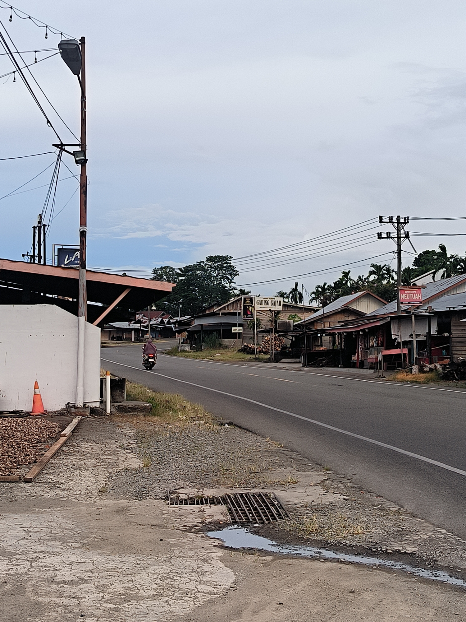 This image captures a roadside scene in Sukamulia, Indonesia, under an overcast sky. A two-lane paved road, marked with a single white line, extends into the distance. On the left, a cracked concrete shoulder features a metal drain grate with a small puddle, and an orange traffic cone. Behind this, a low white wall fronts a building with an exposed rafter roof, and a large utility pole heavily laden with wires and streetlights.
A person on a red motorcycle, wearing a head covering, is seen riding away from the viewer along the road. On the right side of the road, a series of modest, single-story buildings, constructed from wood and possibly concrete, line the street. These structures appear to be homes or small businesses. Another utility pole with multiple power lines stands further down on the right.
Lush green trees, including palm trees, are visible behind the buildings and in the background. Signs with visible text include "LA" on a black banner to the left, and further down the road on the right, "GADING GAJAH" and "MEUTUAH" are displayed on storefront signs. The overall atmosphere suggests a quiet, everyday moment in a semi-urban or rural setting.