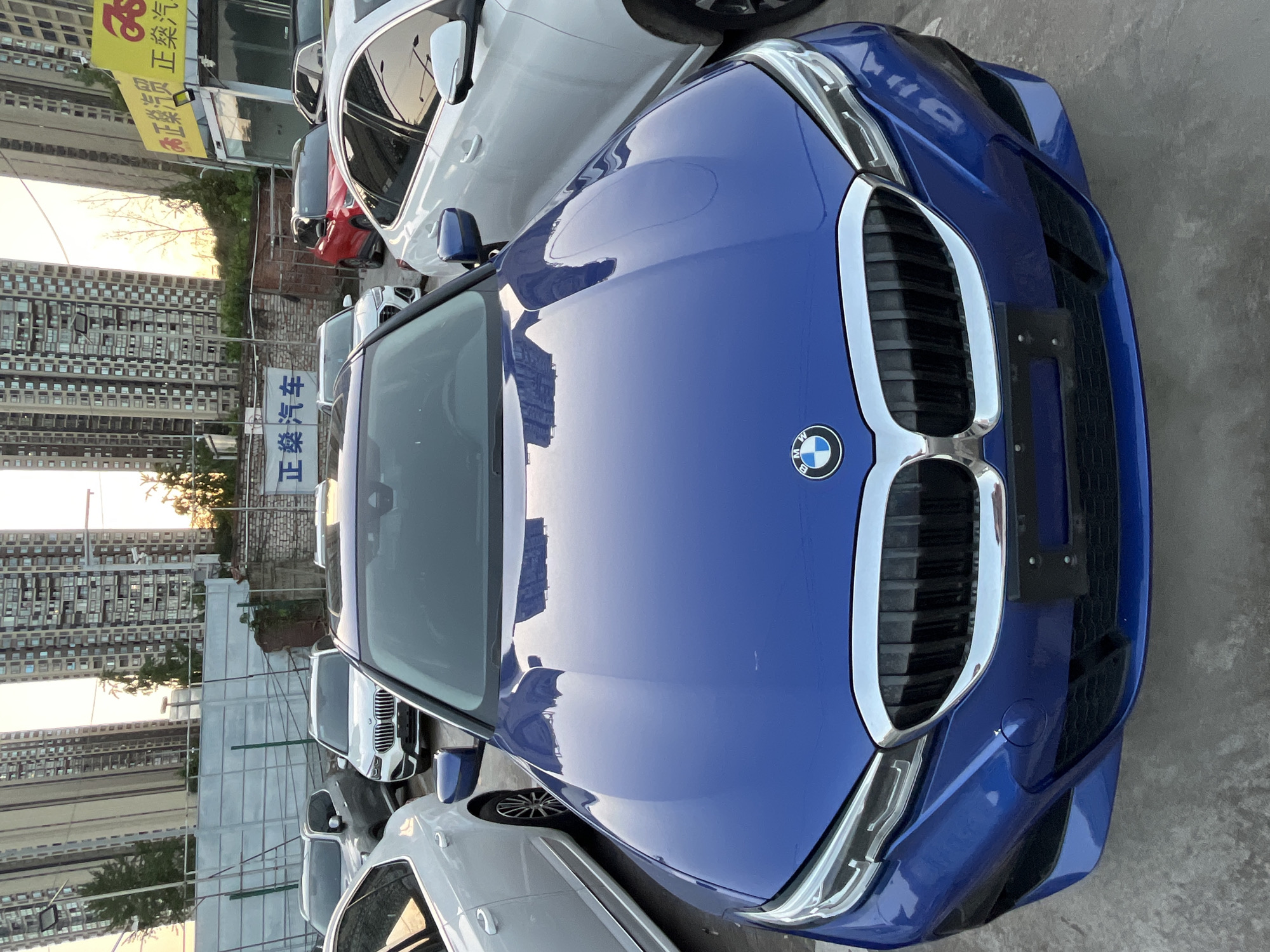 The image shows a blue BMW car parked among other vehicles, possibly in a car dealership lot. The hood of the BMW reflects the surrounding cityscape. The location is Banan, China, indicated by signage in Chinese characters visible in the background. The environment includes buildings and other cars.