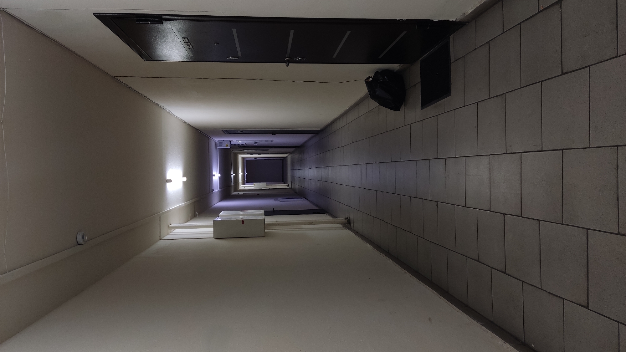 An upward-angled view reveals a long, narrow corridor within a building. The corridor is dimly lit, with light fixtures spaced along the left wall. The right wall features a tiled surface. At the near end of the corridor, a dark-colored door is visible above. Further down the corridor, an object resembling a black bag hangs near another door. The far end of the corridor appears dark. A white object sits on the floor along the left side. The location of this image is unspecified, and information about Krasnodar, Russia, is not evident within the visual content.