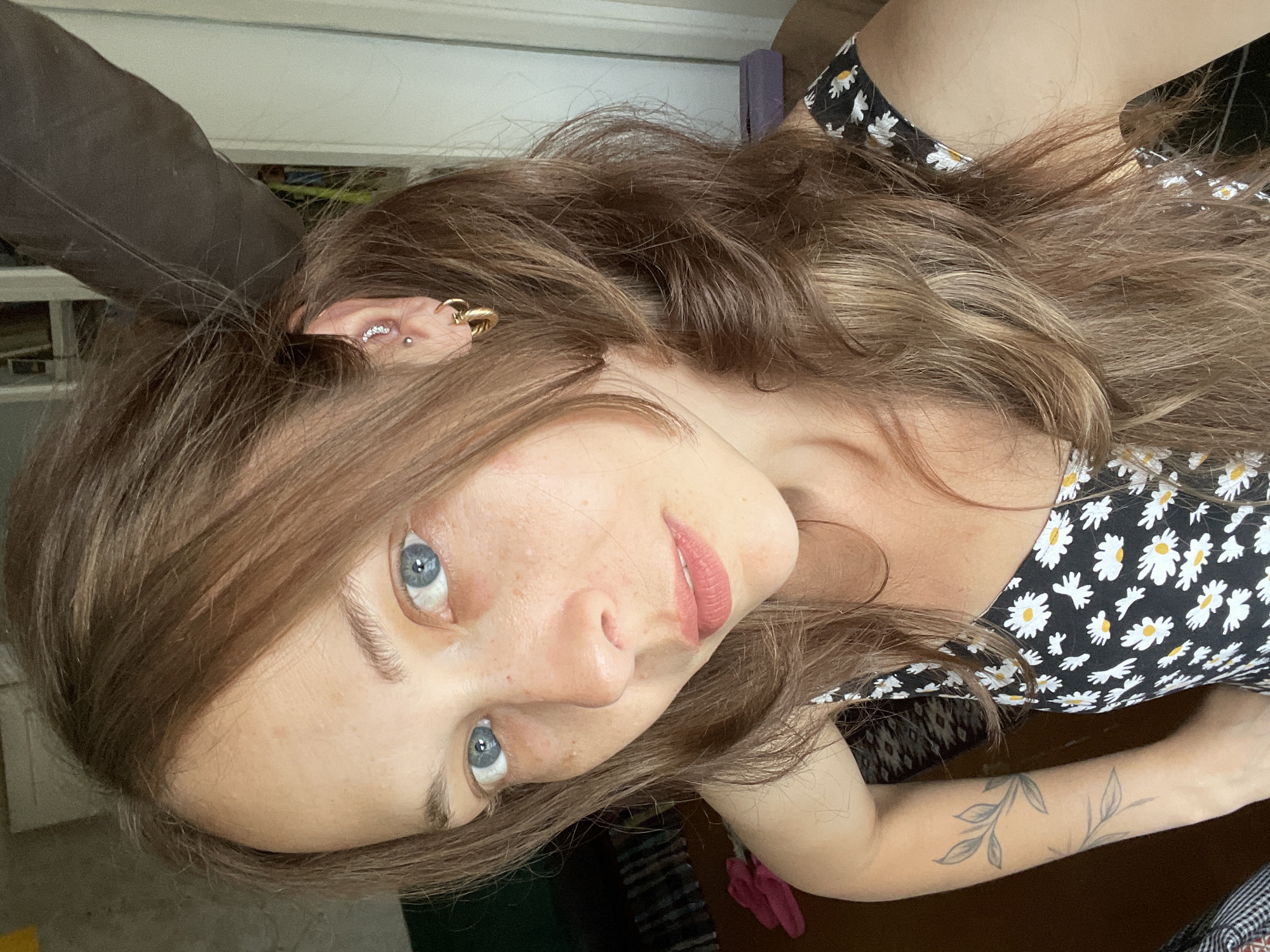 A close-up, selfie-style shot shows a young Caucasian woman with long, wavy, light brown hair and blue eyes. She has freckles on her nose and cheeks. She is wearing a black dress with a daisy print. She has a large feather in her hair, and multiple piercings in her left ear. She also has a tattoo of a plant stem on her left arm. The background is blurry and appears to be indoors, with a wooden surface visible in the foreground.