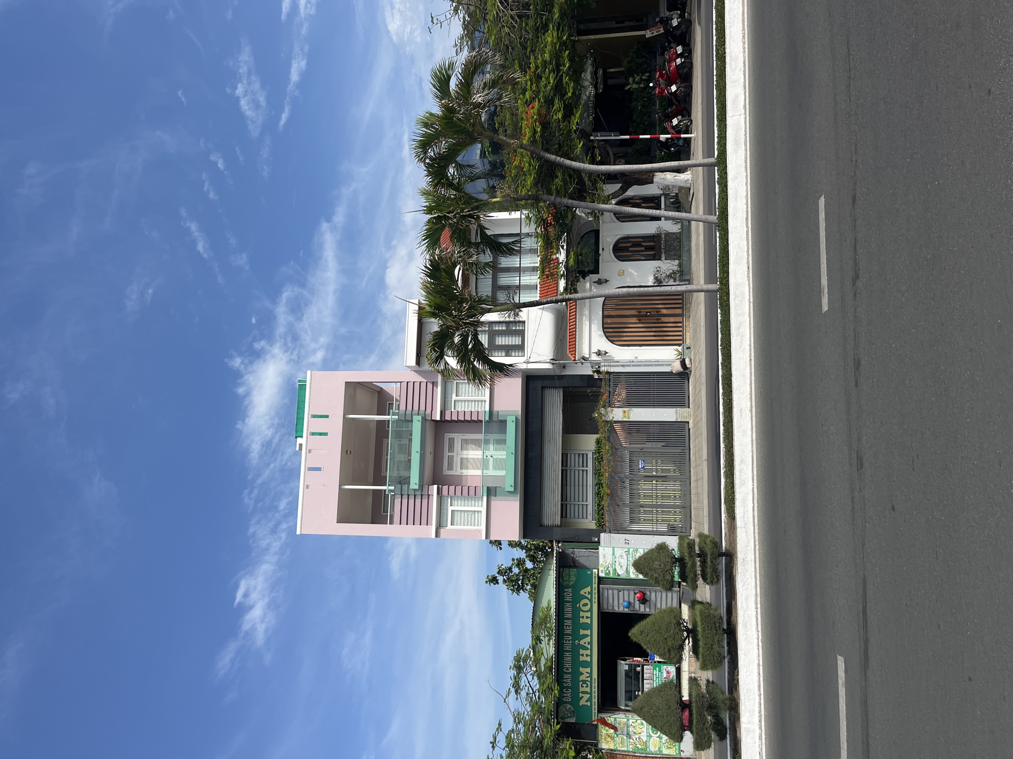 The image shows a street scene in Bac Nha Trang Ward, Vietnam. A multi-story pink building is visible on the left side of the image. Adjacent to this building is another building with a white façade and a red tiled roof. In front of the buildings are palm trees. A business establishment with a green signboard is also visible, displaying Vietnamese text. A paved road with a white line is present on the right side of the image. Red motorbikes are parked near the curb. The sky is blue with sparse clouds.