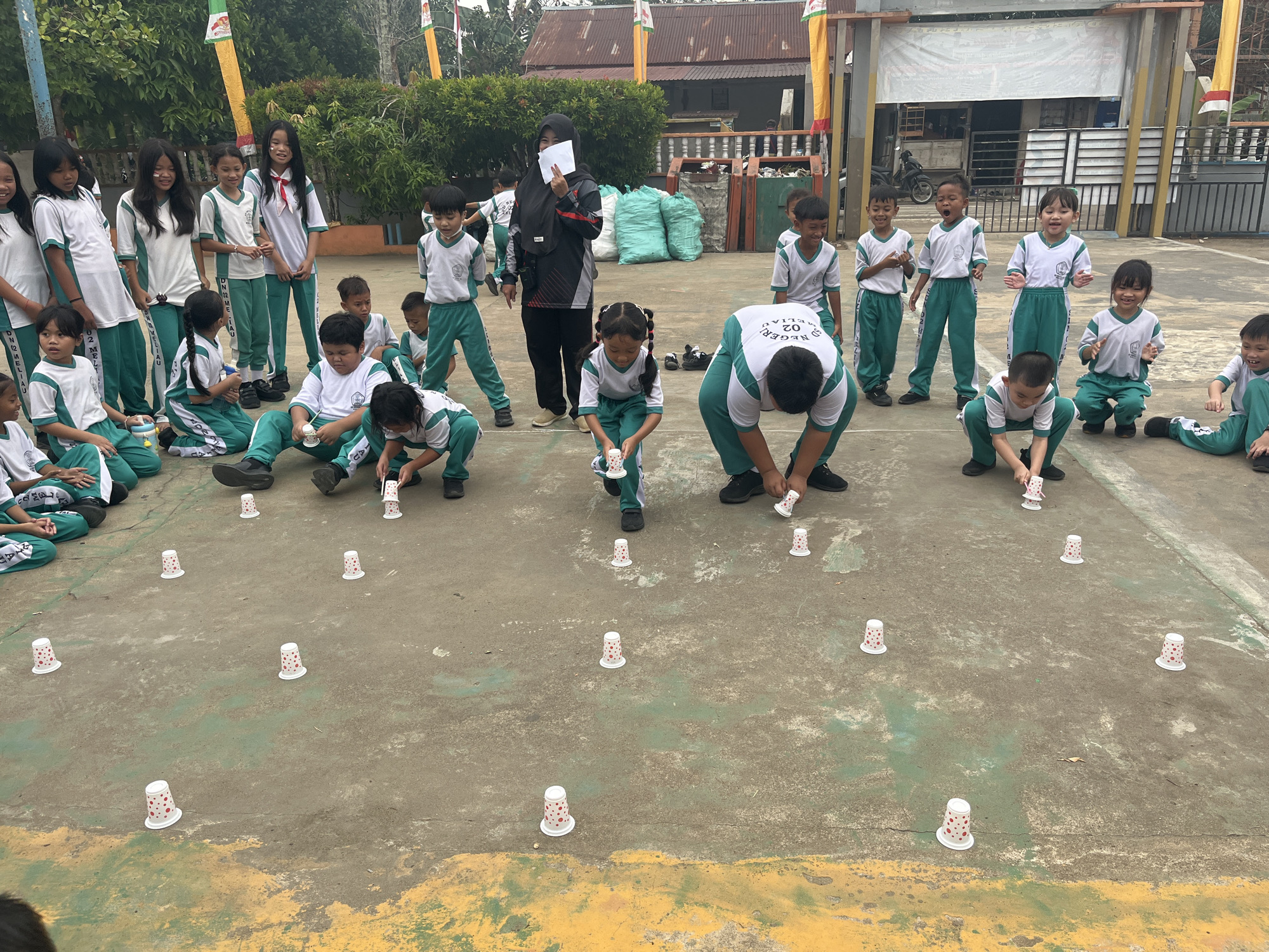 In Meliau Hulu, Indonesia, a group of children in matching green and white uniforms are engaged in an outdoor activity on a paved court. The scene appears to be in a schoolyard, with a building and trees visible in the background. Several children are seated on the ground, while others are standing or crouching. They are participating in a game involving small, white paper cups with red polka dots, which are scattered across the court. Some children are shown reaching for or interacting with the cups. A woman in a dark hijab and grey jacket is standing and holding a piece of paper, seemingly observing or instructing the children. The ground is a worn, mottled surface, with painted lines in yellow and green. The overall atmosphere suggests a lively and organized event for the children.