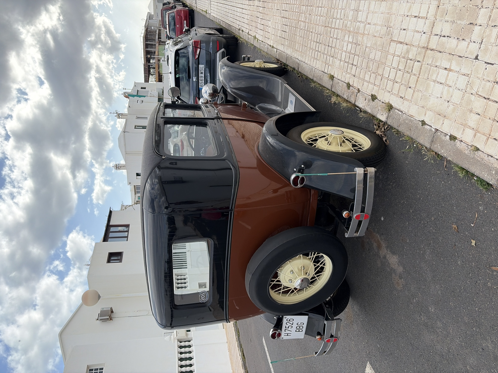 The image captures a street scene in Tías, Spain, under a partially cloudy sky during daytime. The primary subject is a classic, two-tone car, dark brown on the lower body and black on the upper body and fenders, parked on the right side of an asphalt road. It features cream-colored spoke wheels, with a spare tire mounted at the rear. Chrome bumpers are visible at the front and rear of the car, along with red taillights. The rear license plate reads "H 7526 BBG". A small "AVA" sticker is visible on the rear window.
Further down the street, several modern cars are parked in a line. One dark grey SUV-like car is clearly visible, with its license plate reading "6569 MSL" and the text "arona" (likely referring to the SEAT Arona model) beneath it. The vehicles are parked alongside a sidewalk paved with light-colored, square tiles, with sparse grass and weeds growing in the cracks and along the curb.
On the left side of the street, a row of white buildings with simple, modern architecture can be seen, featuring dark-framed windows and a few exterior elements like a spherical light fixture and an air conditioning unit. The sky above is bright blue with scattered, fluffy white clouds, indicating clear weather. The overall scene depicts a quiet street in a residential or commercial area.